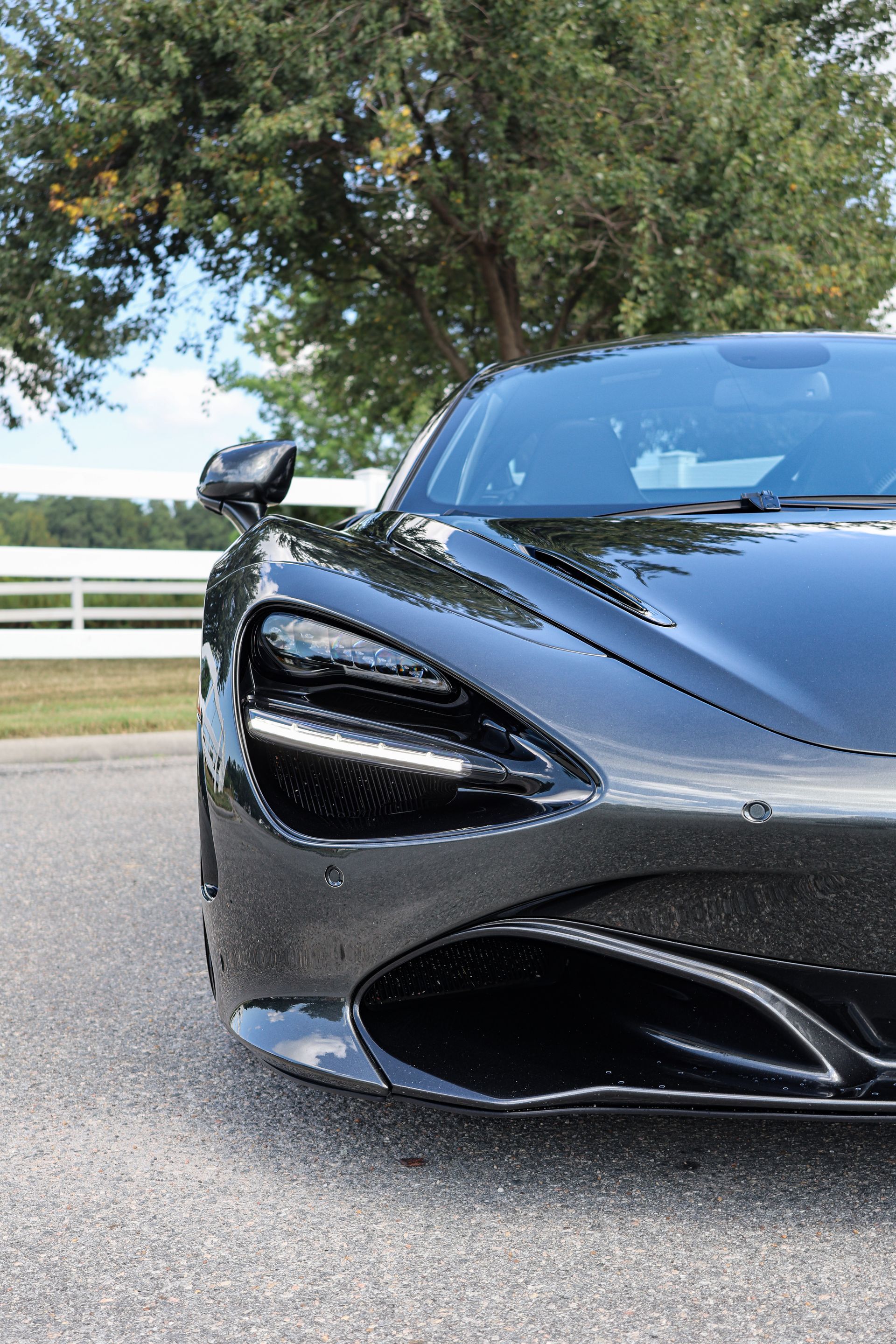 Close-up of a sleek, black McLaren 720S sports car parked on an asphalt road in front of a white fence and green trees.