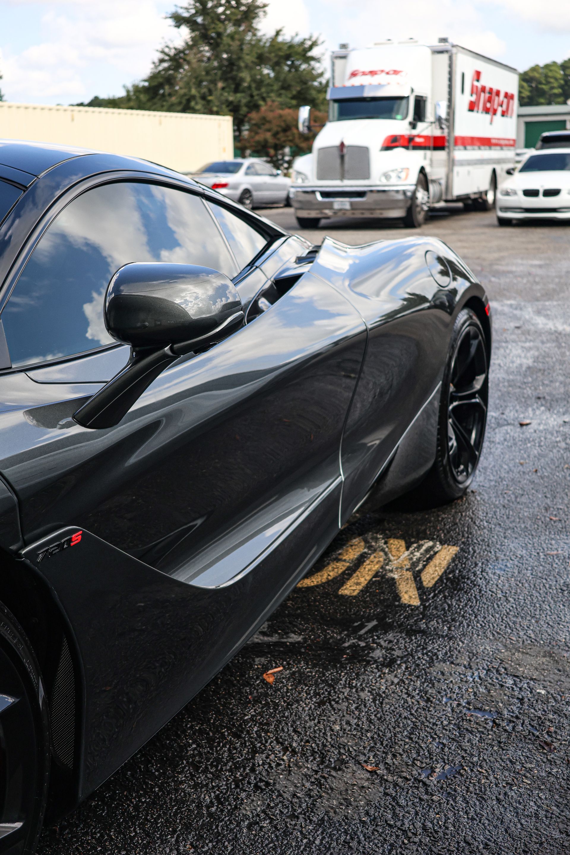 Dark gray sports car parked, Snap-on truck and other vehicles in the background. Wet pavement.