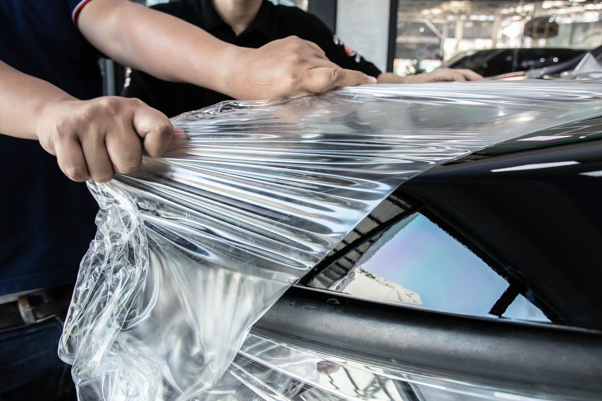 Person stretching clear vinyl wrap over a car hood in an auto shop