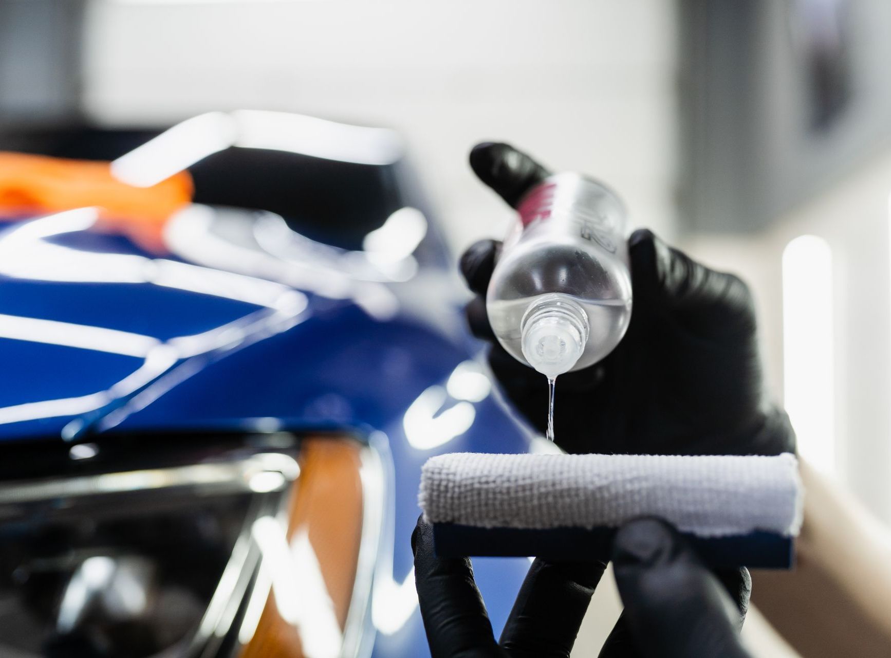 Gloved hands applying liquid to a car's blue surface using a sponge applicator.