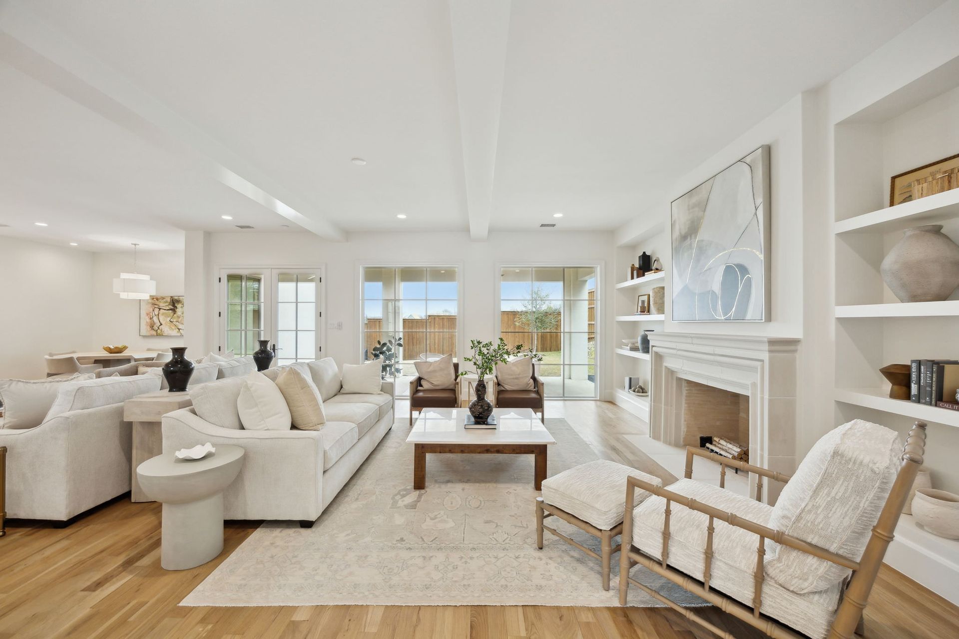 Bright living room with white sofas, fireplace, and built-in shelves, with a view of a backyard through glass doors.