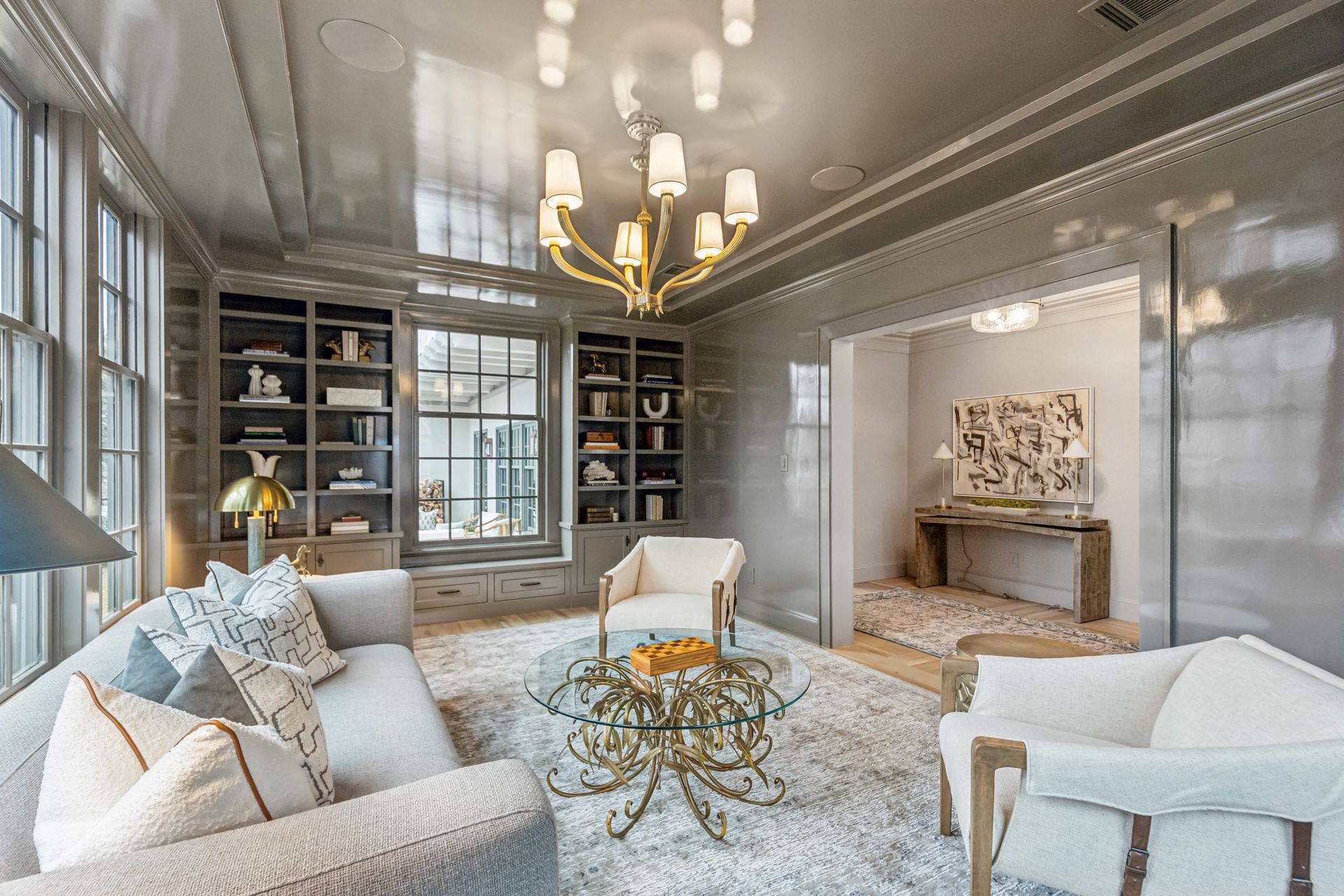 Elegant living room with light gray walls, bookshelves, and seating. A gold chandelier hangs overhead.