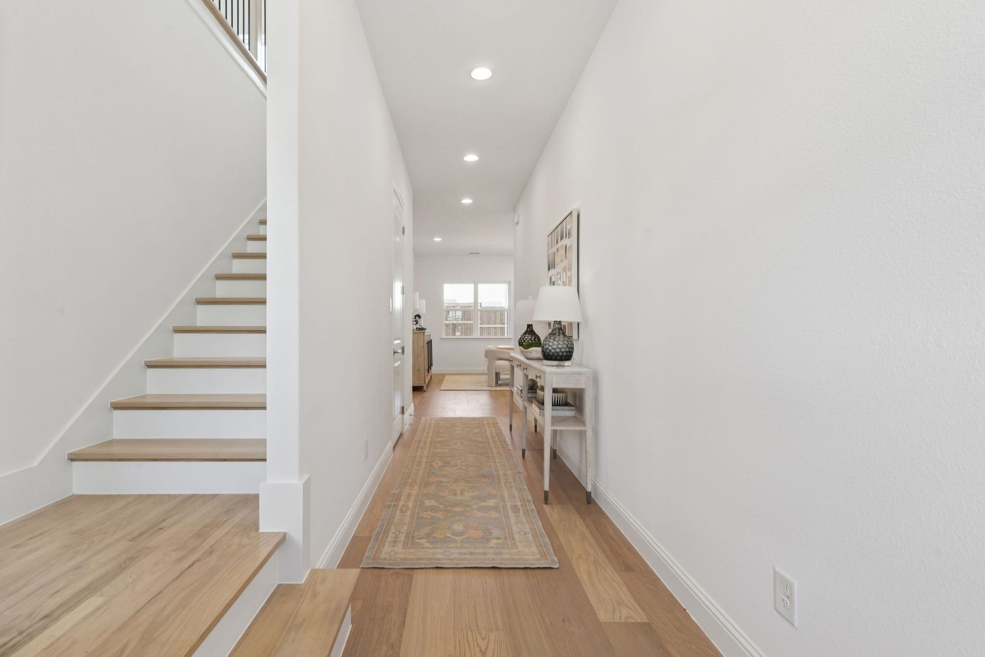 Bright hallway with stairs, light wood floors, and white walls. Beige rug and console table.