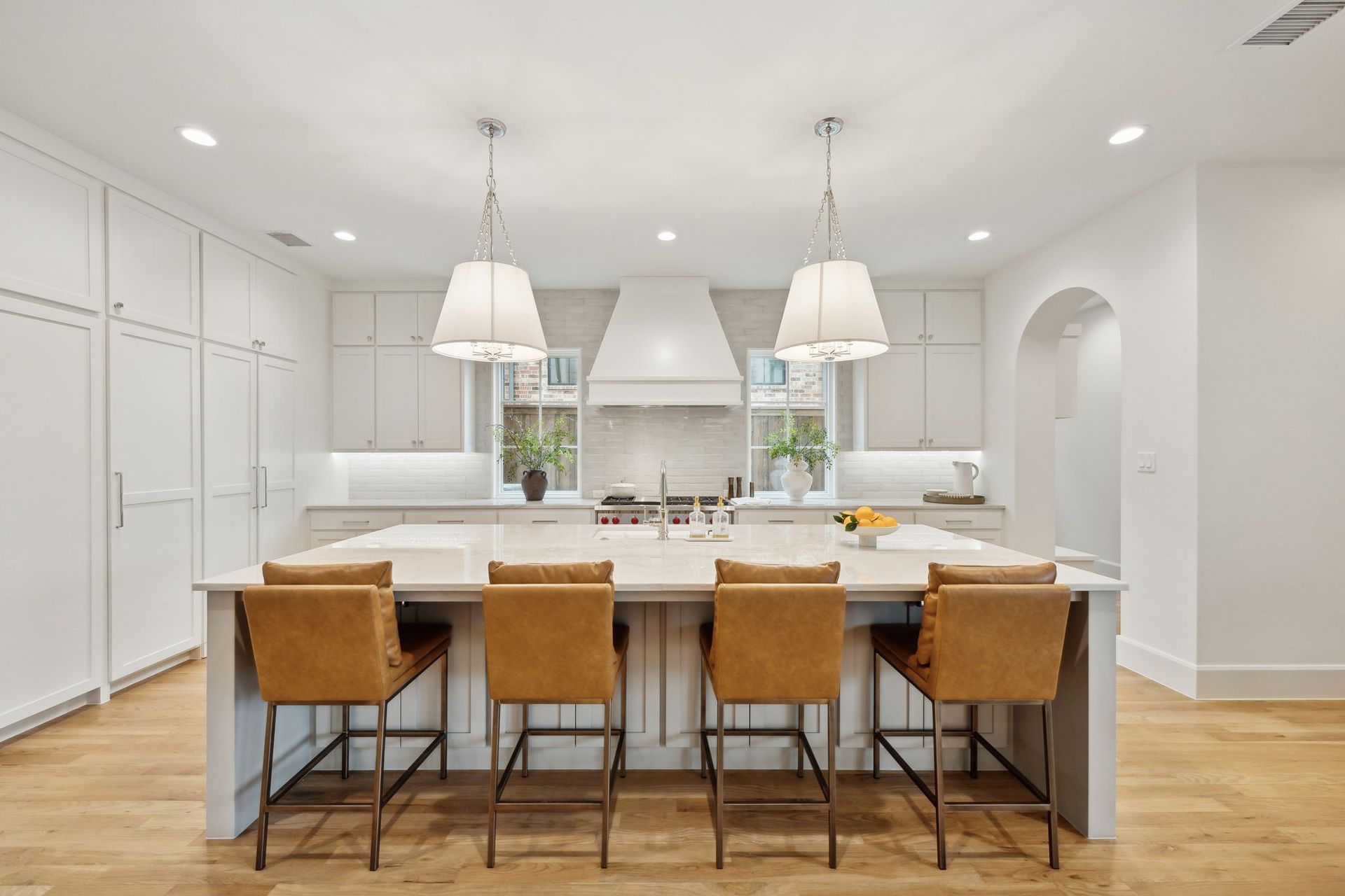 Bright, white kitchen with large island, pendant lights, brown leather bar stools, and arched doorway.