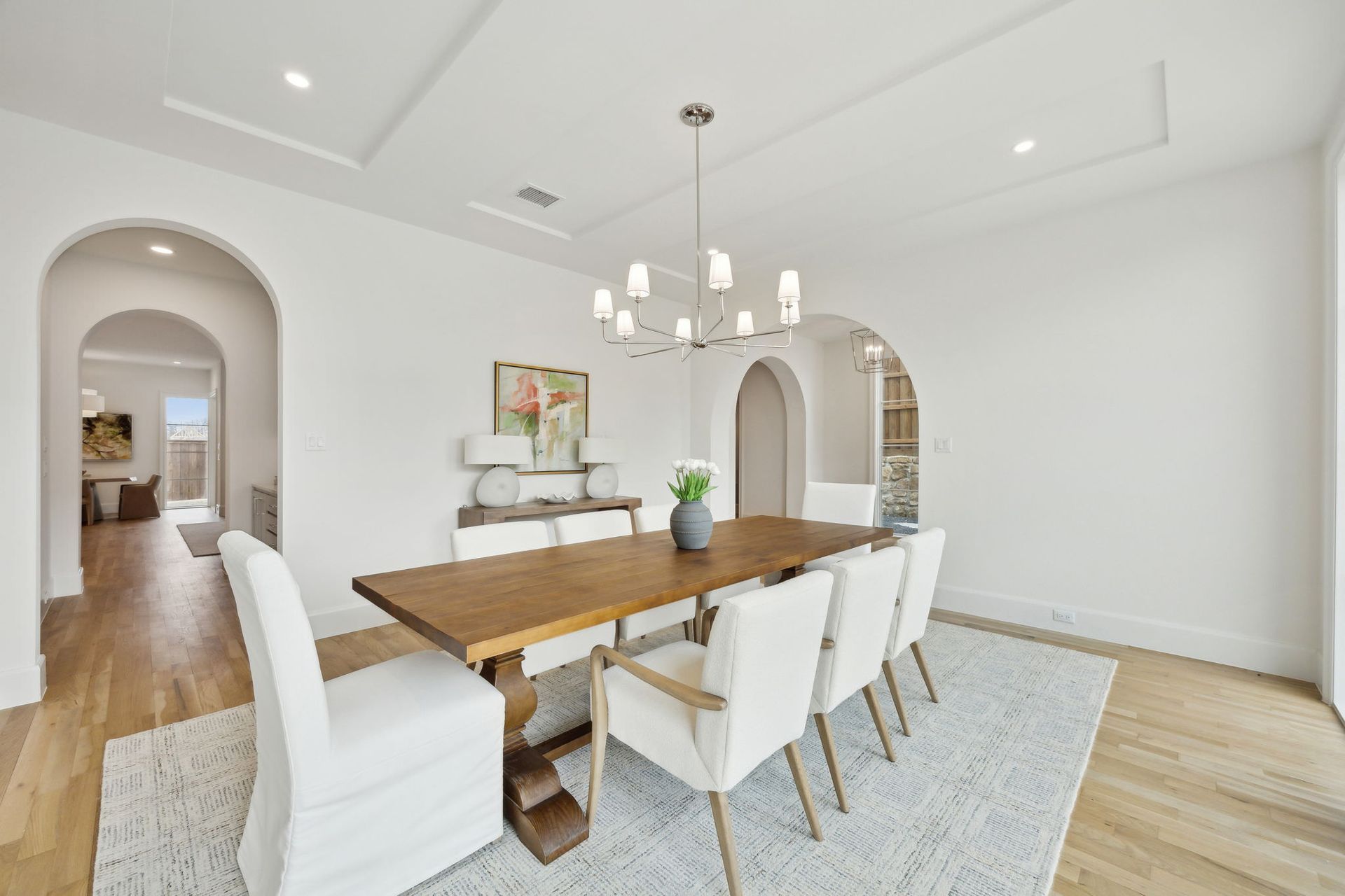 Dining room with wooden table, white chairs, and modern chandelier on a white rug.