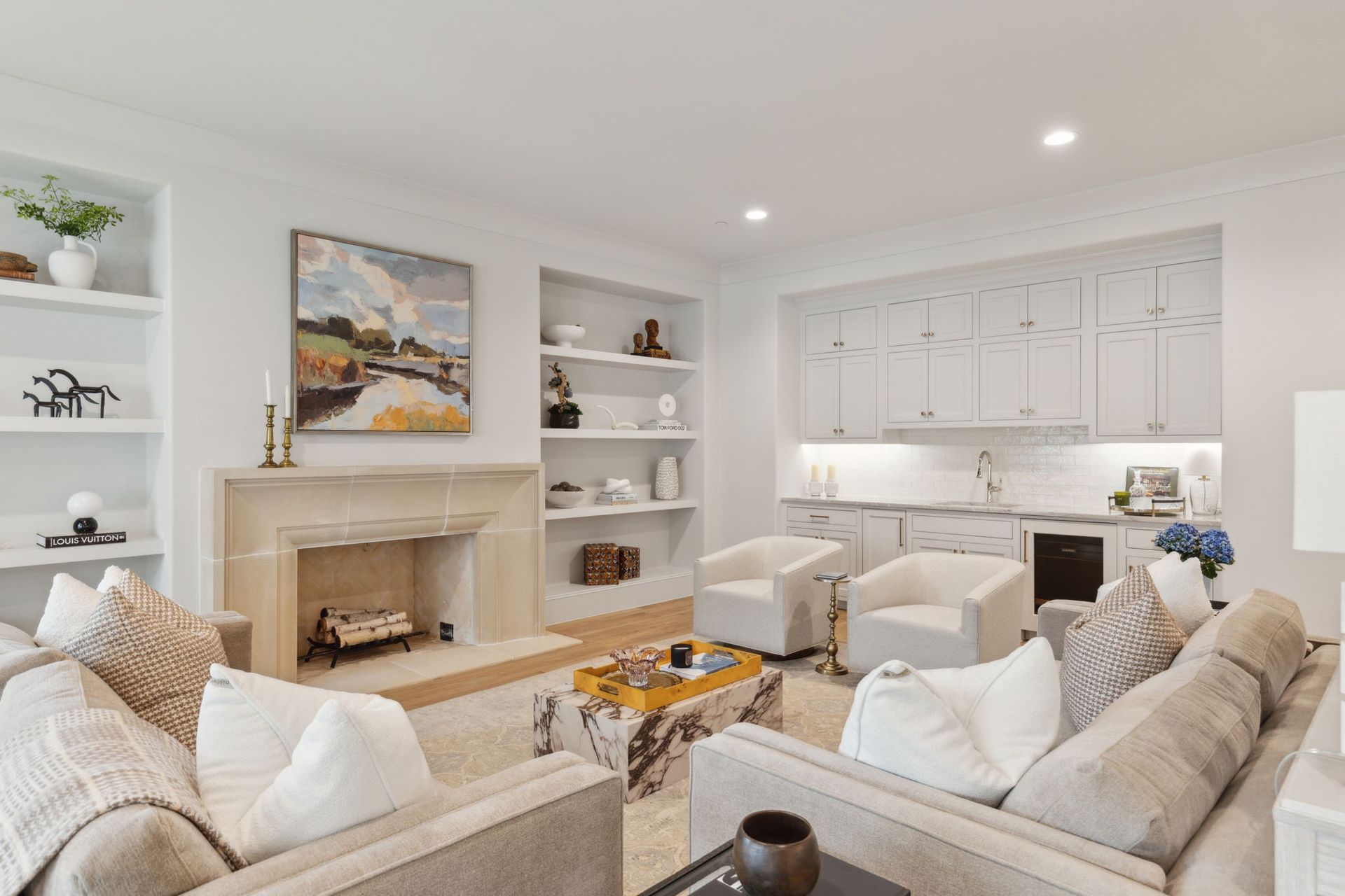 Living room with fireplace, built-in shelves, and a small kitchen area; white walls, neutral furniture.