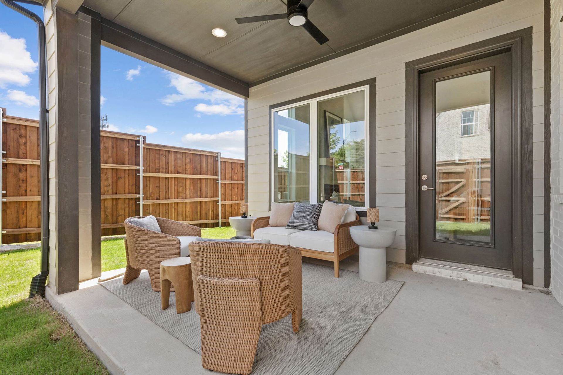 Patio with wicker furniture, rug, and a door, beneath a shaded roof, against a wooden fence.