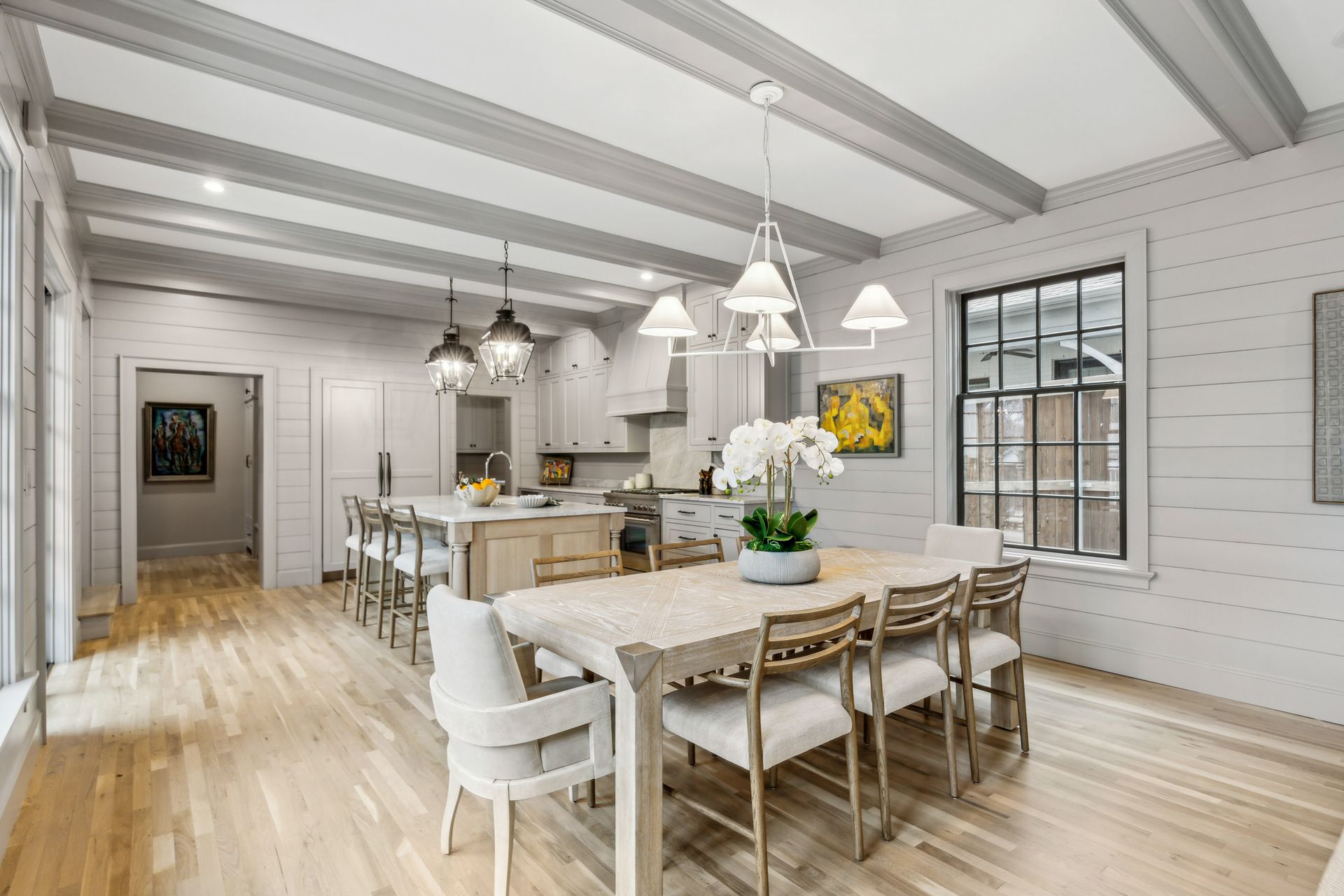 Open-concept kitchen and dining room with light wood floors, white walls, and gray beams.