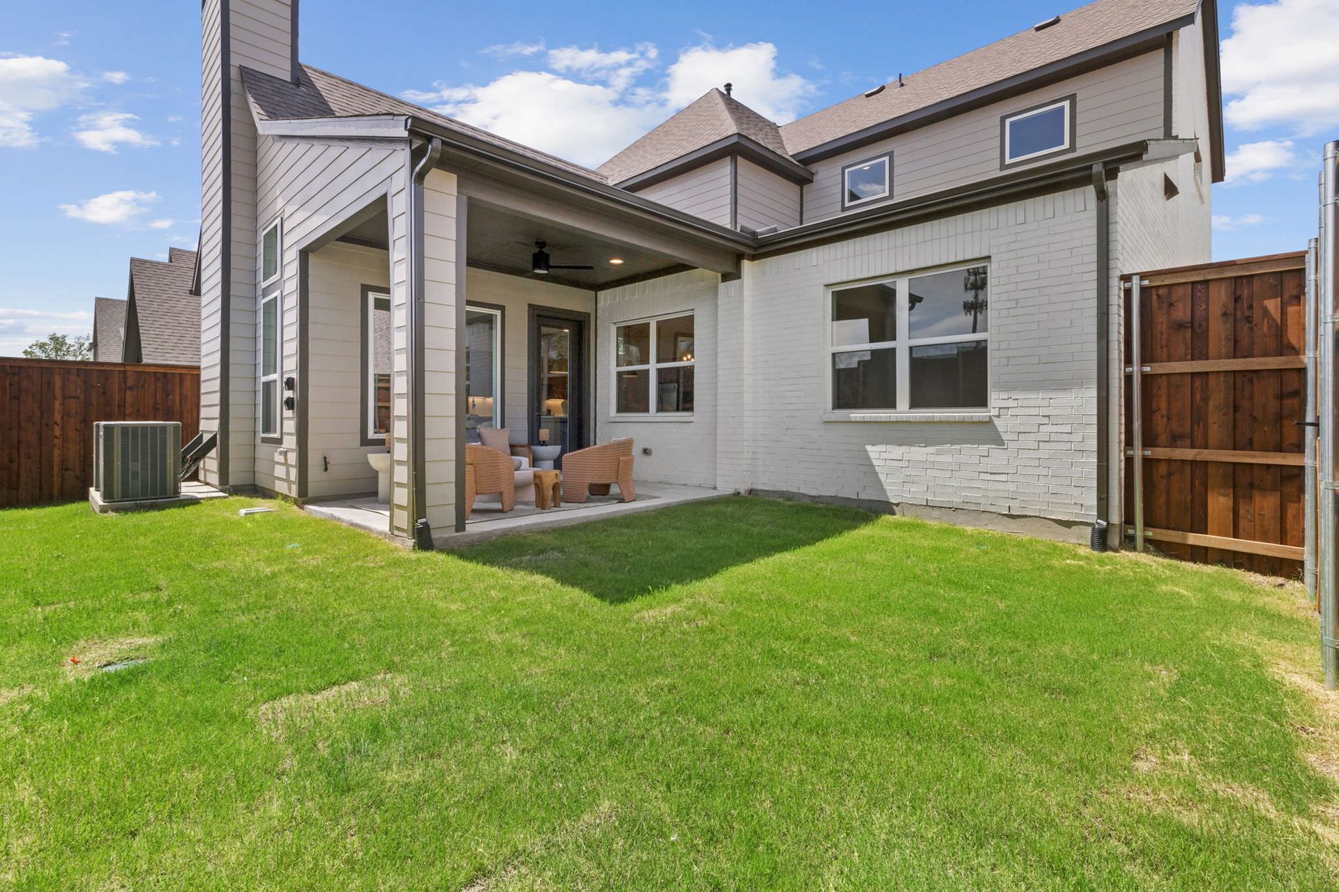 Backyard with green grass, patio with chairs, brick building and wooden fence.