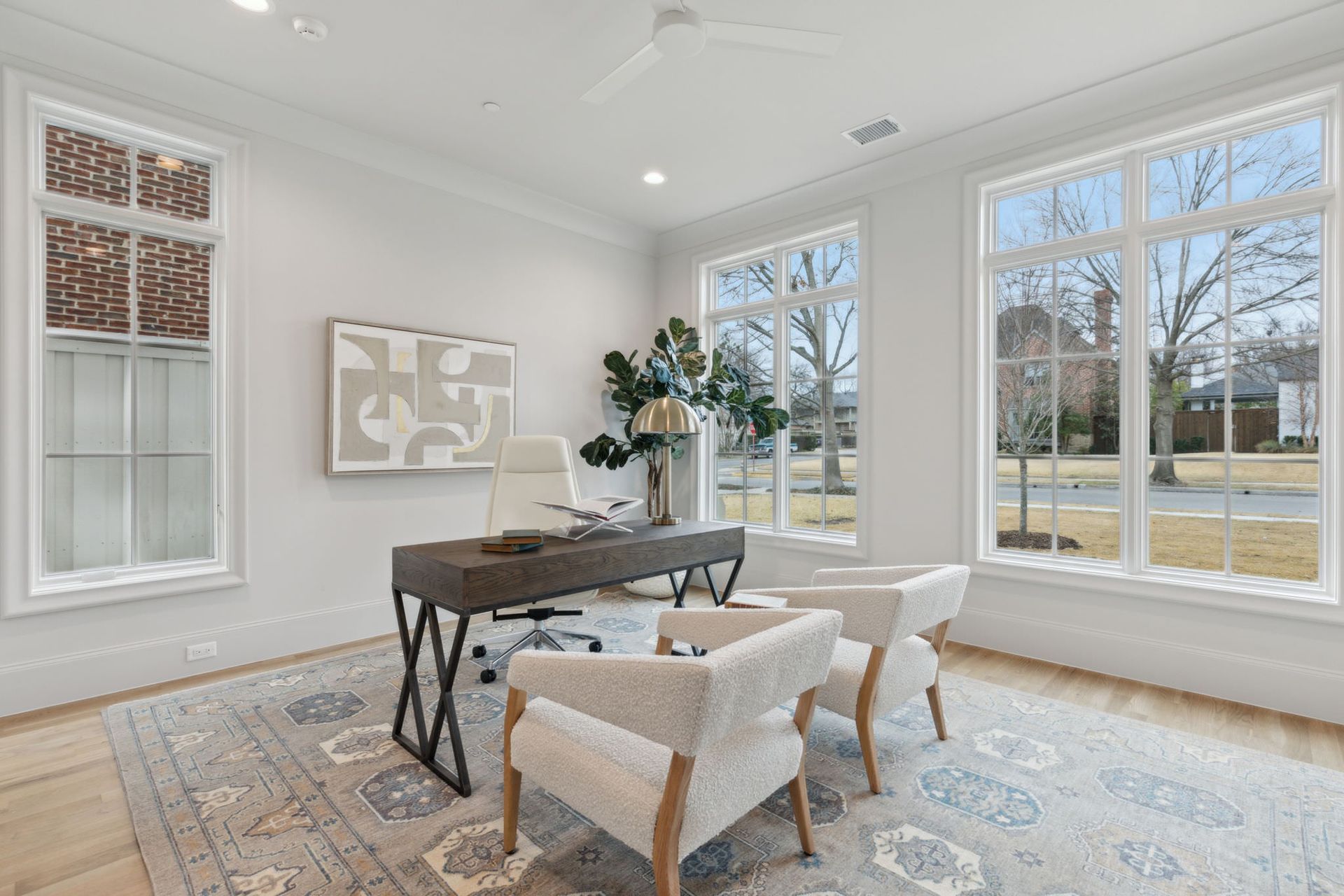Home office with desk, chairs, and large windows. Neutral color scheme, rug, and artwork.