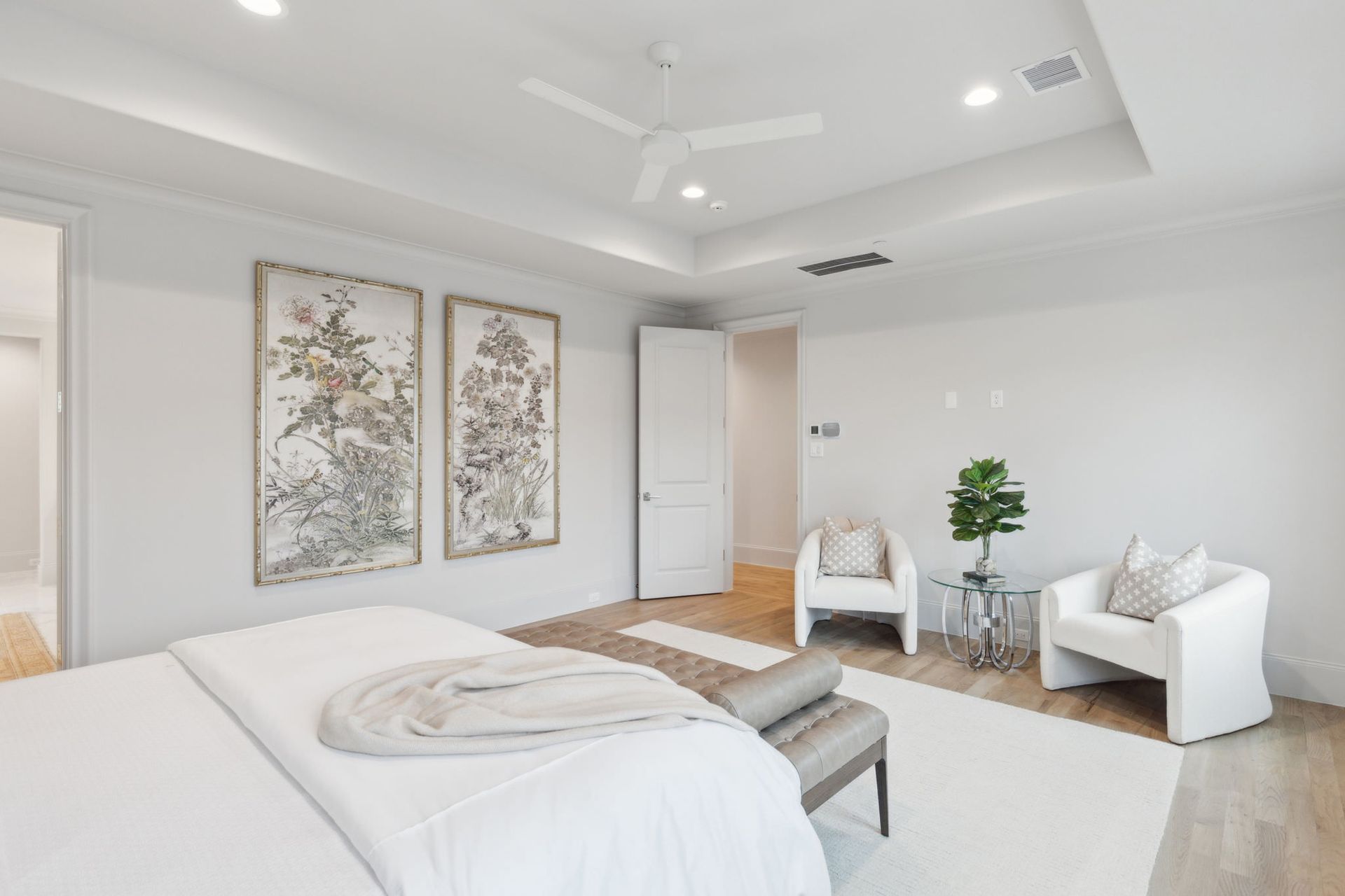 Spacious, bright bedroom with white walls, artwork, seating area, and light wood flooring.