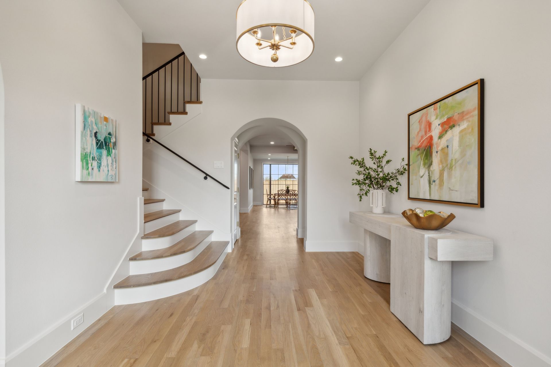 A hallway with hardwood floors , stairs , a table and a painting on the wall.