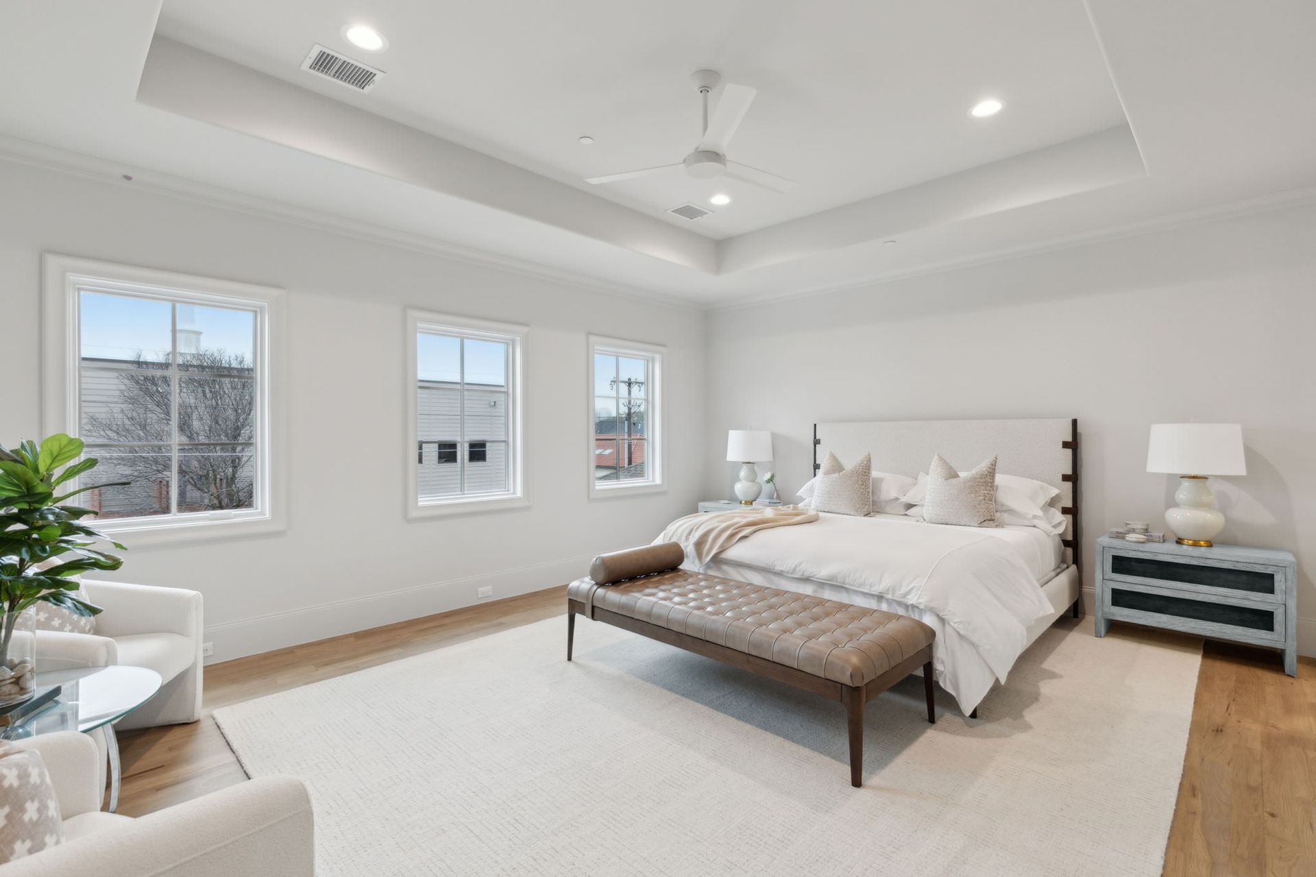 Spacious, white bedroom with windows, king bed, seating area, and a long brown bench at the foot of the bed.