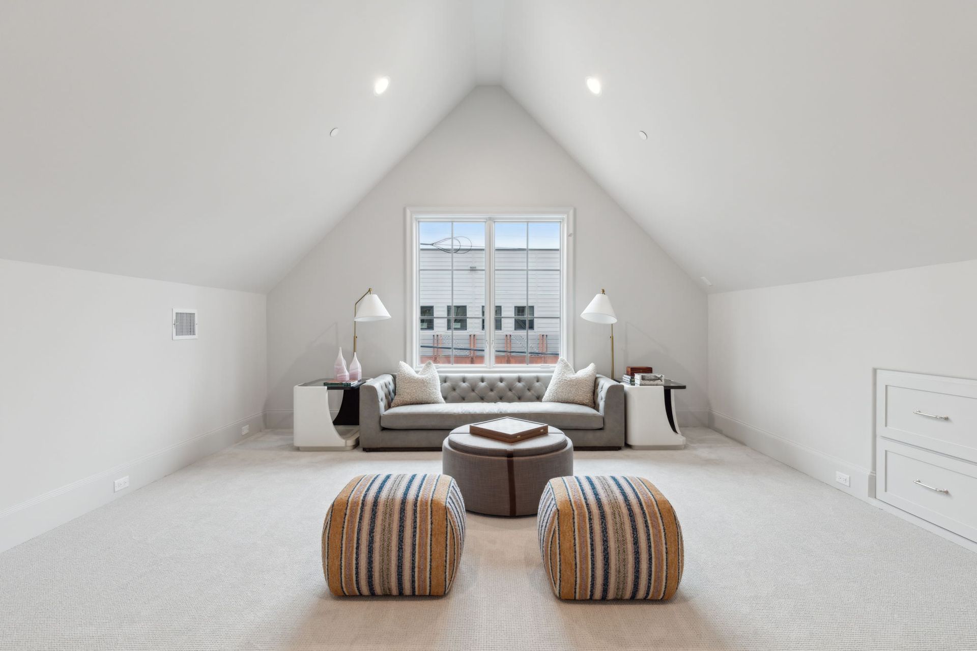 Spacious, bright attic room with a gray sofa, ottomans, and a window.