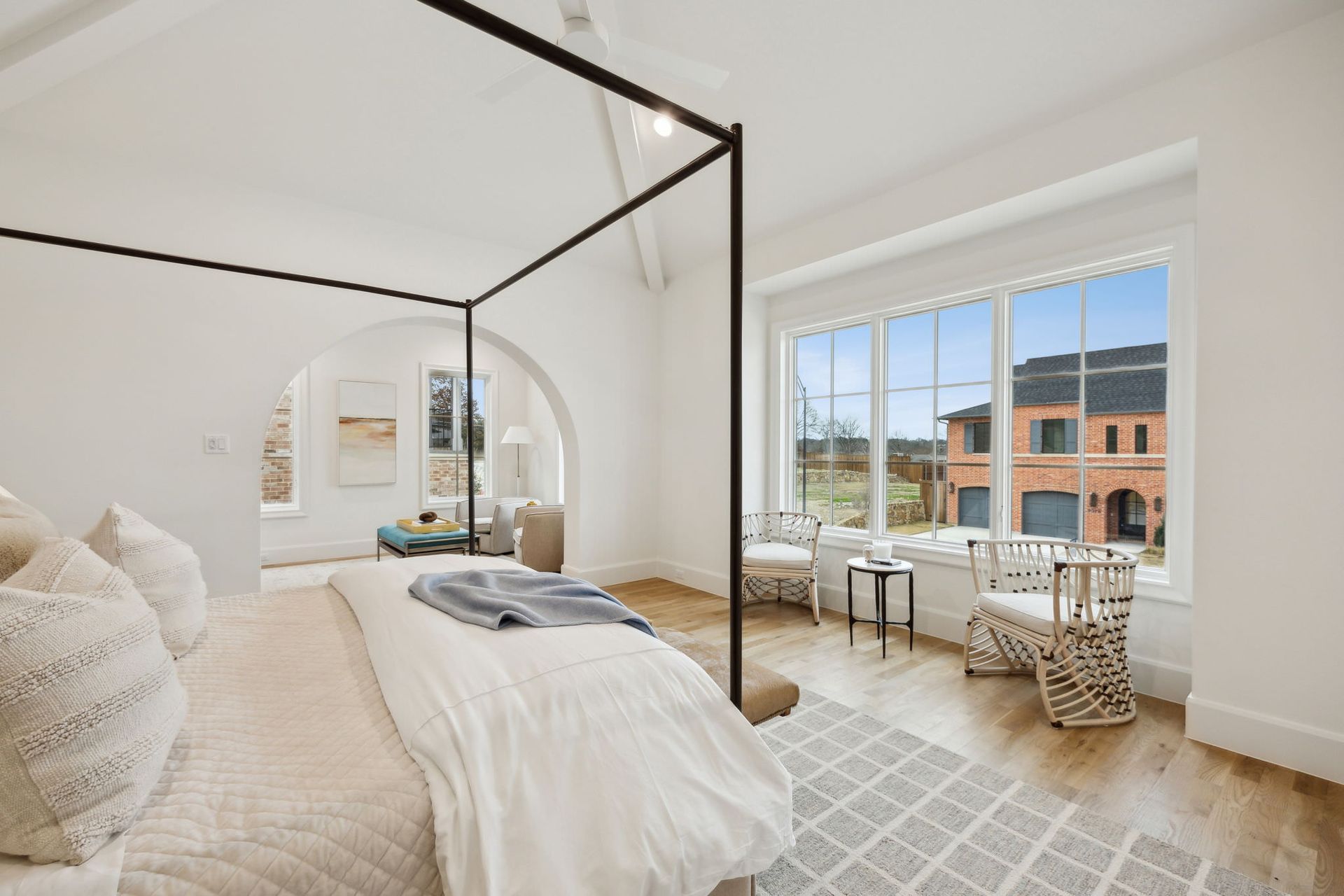 Bedroom with four-poster bed, window seating, and archway to a seating area, white walls and light wood floors.