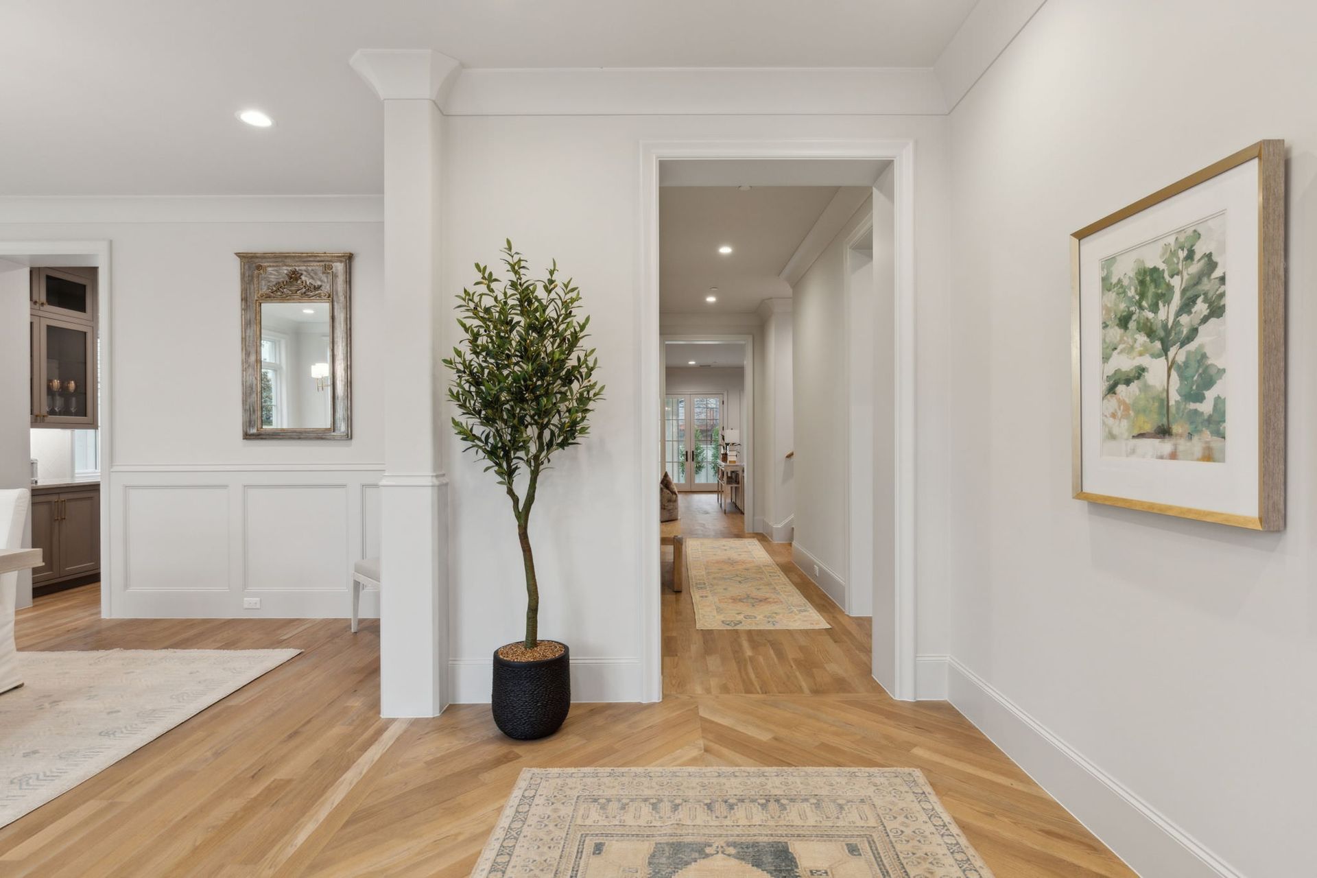 Bright entry hall with hardwood floors, potted tree, artwork, and a hallway leading to the back.