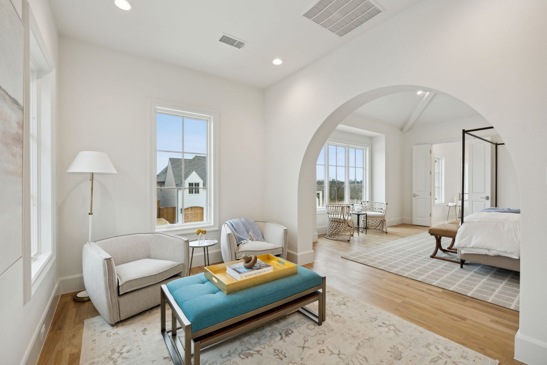 Bright white bedroom with arched doorway. Two white chairs, teal ottoman, and a view of the outside.