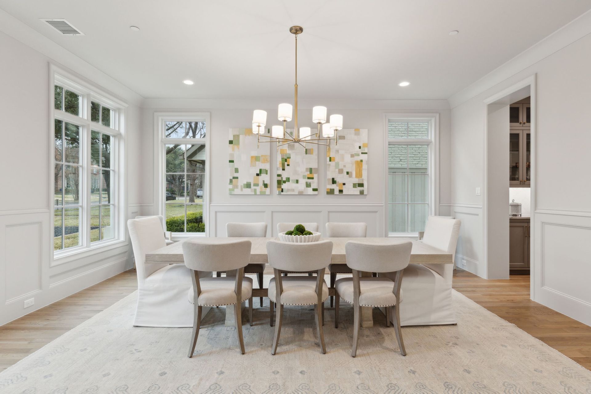 Elegant dining room with table, chairs, rug, chandelier, and framed art; white walls, light wood floor.