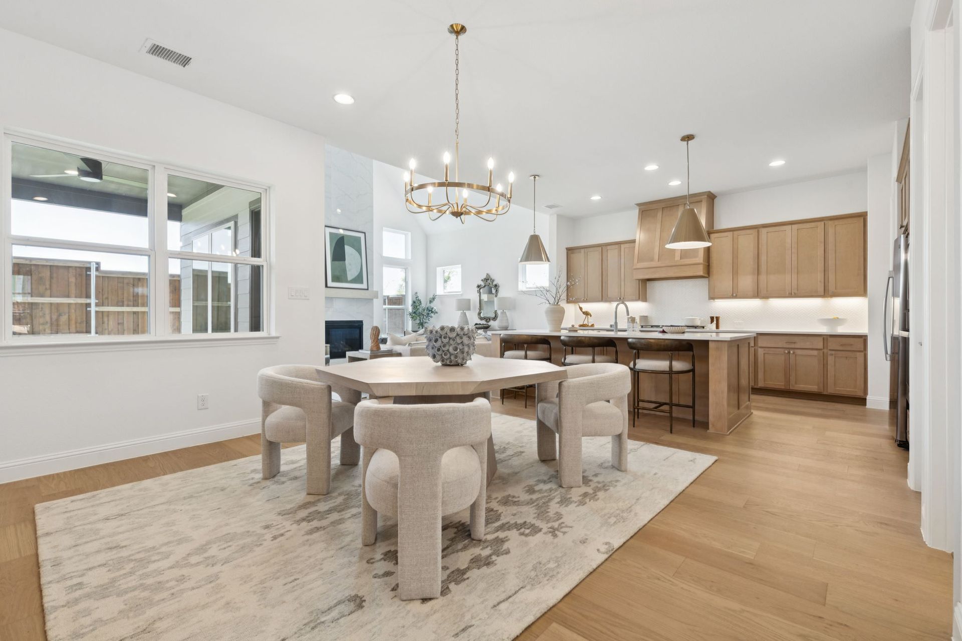 Dining room with round table, chairs, rug, and kitchen in background. Bright and airy interior, light wood floors.