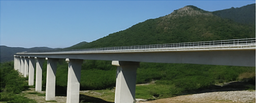 Un lungo ponte di cemento che attraversa una valle, con verdi colline sullo sfondo sotto un cielo azzurro.