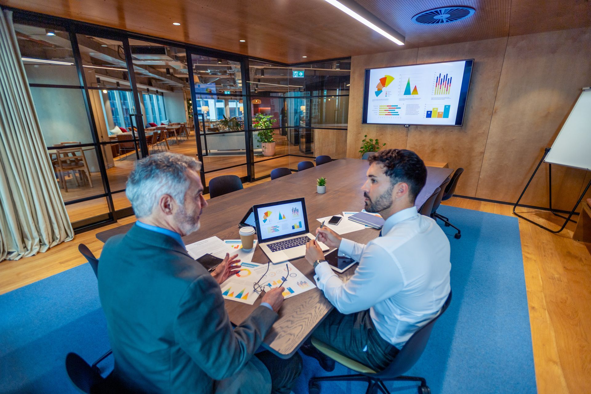 Two men are sitting at a table in a conference room looking at a laptop.