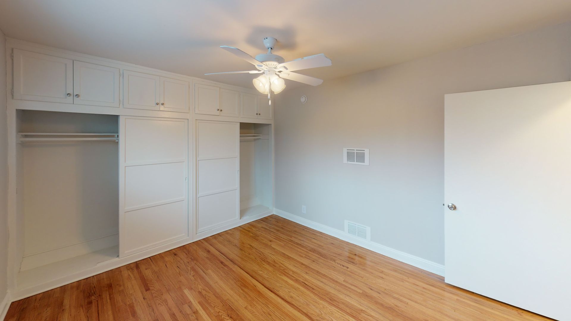 Empty bedroom with wood floor, built-in white cabinets, a ceiling fan, and a closed white door.
