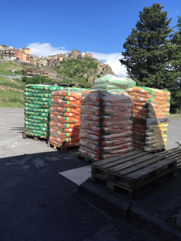 Pallet impilati di sacchi verdi e arancioni in un piazzale all'aperto, con una cittadina di montagna sullo sfondo.