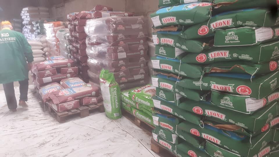 Un operaio con una giacca verde si trova in un corridoio di un magazzino pieno di pile di prodotti insacchettati su pallet di legno.