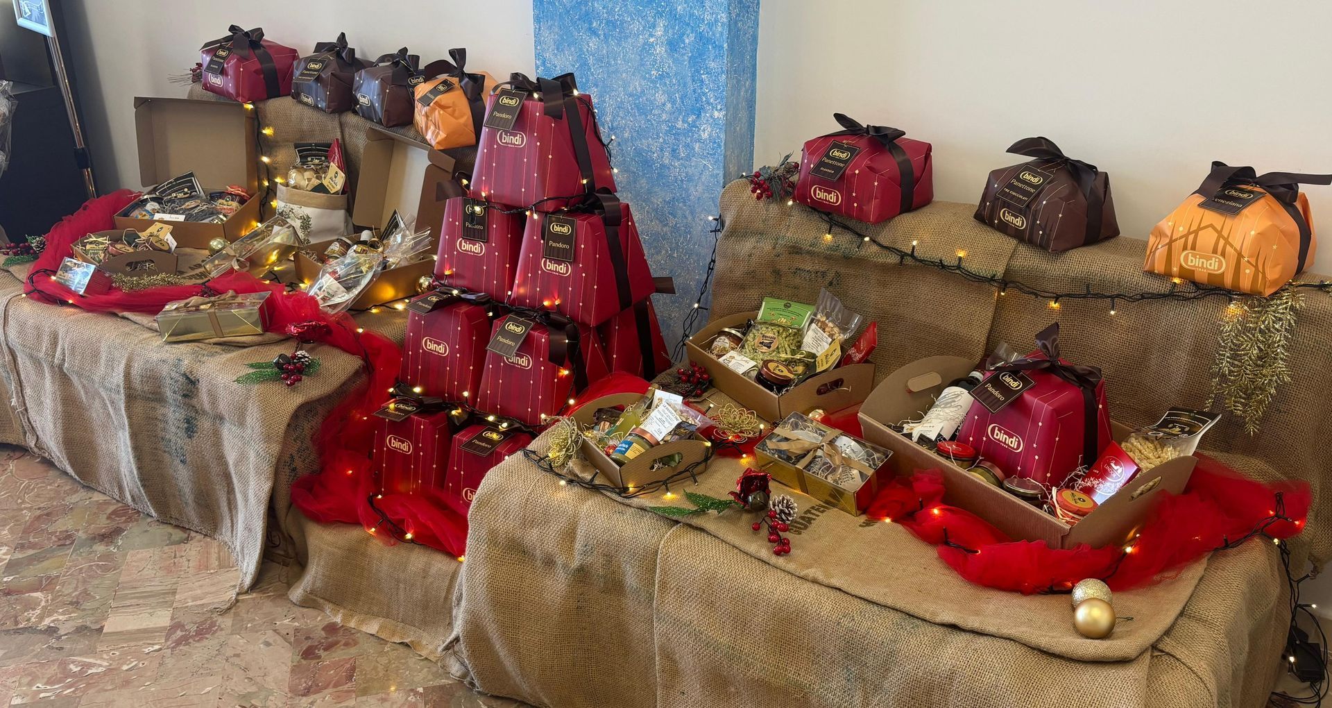 Esposizione di regali natalizi su tavoli ricoperti di tela di iuta, con articoli in scatole rosse, cesti regalo e dettagli dorati.