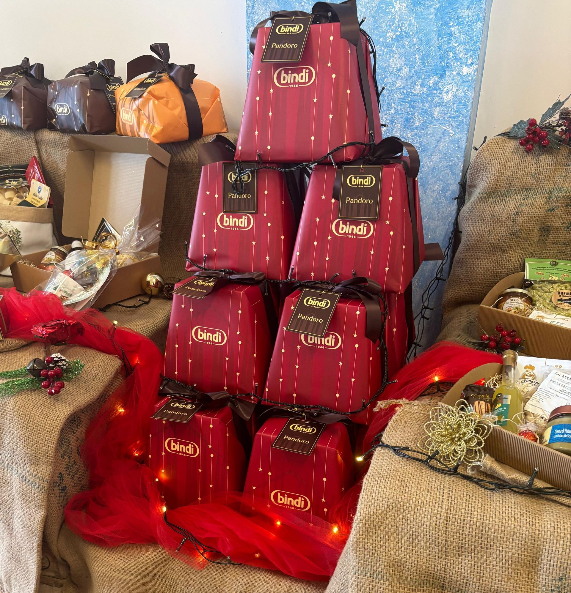 Una composizione piramidale di scatole di panettone rosse del marchio Bindi, decorate con nastri marroni e luci festive su un telo di iuta.