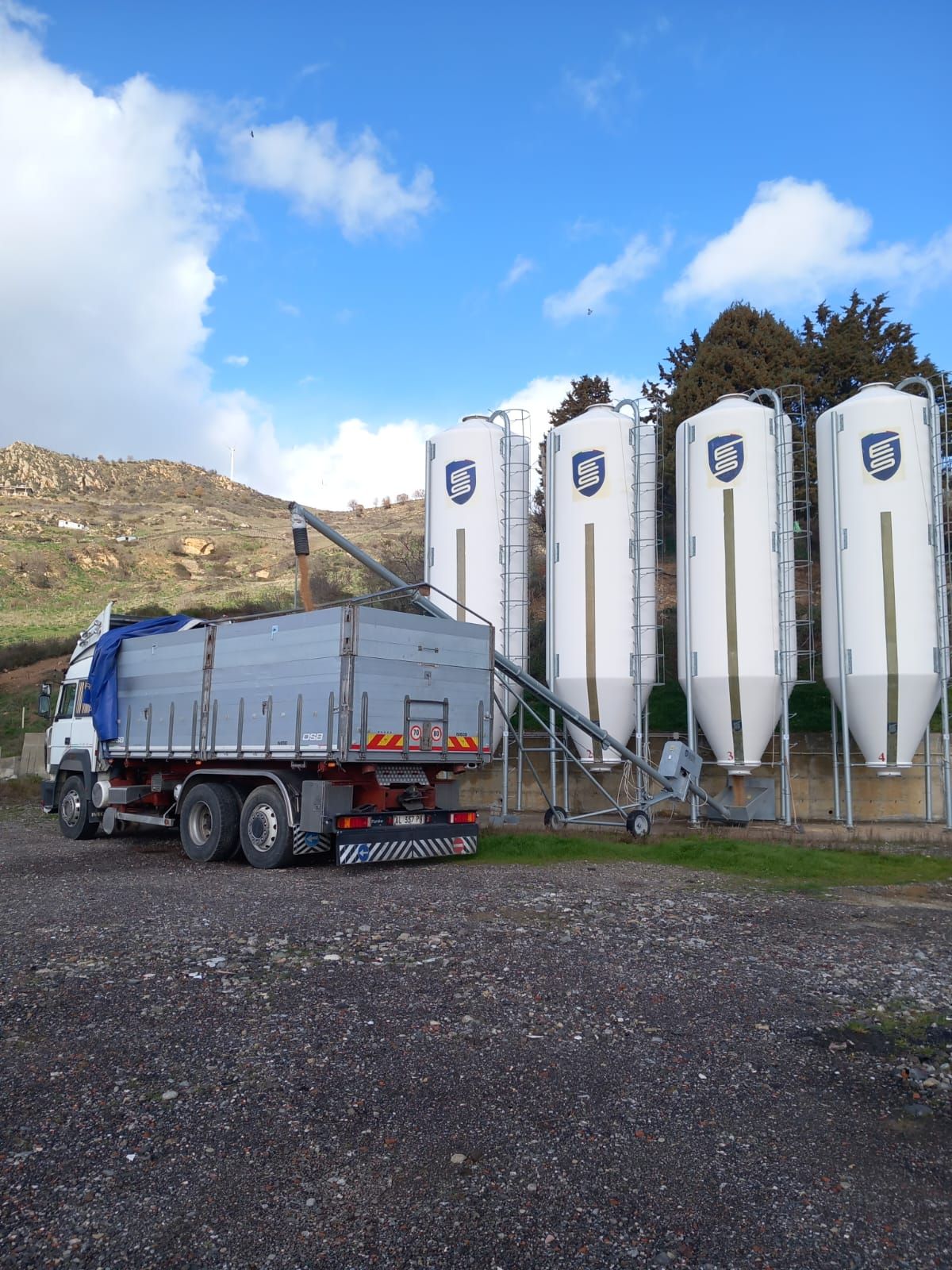 Un camion con un nastro trasportatore allungabile riempie uno dei quattro alti silos industriali bianchi situati su una collina in campagna.