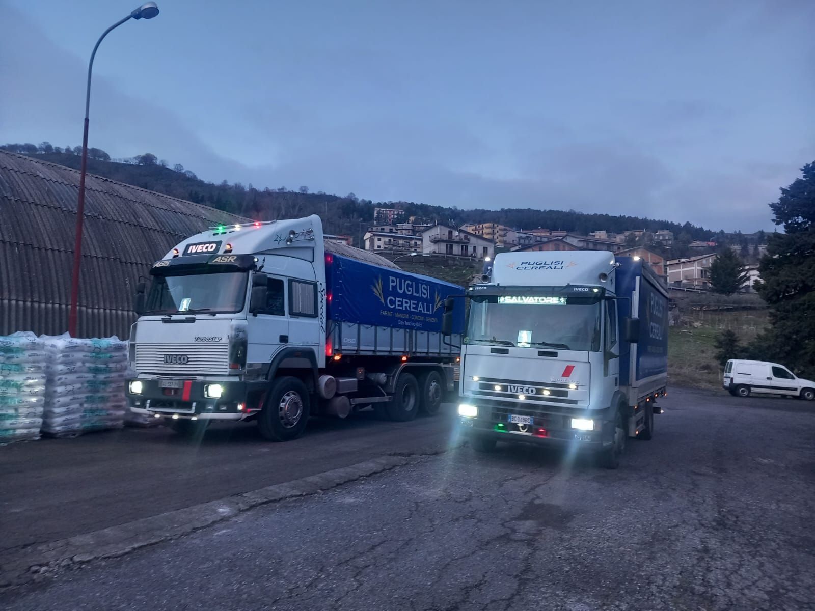 Due camion bianchi parcheggiati fianco a fianco in un parcheggio asfaltato al crepuscolo, con una cittadina collinare sullo sfondo.