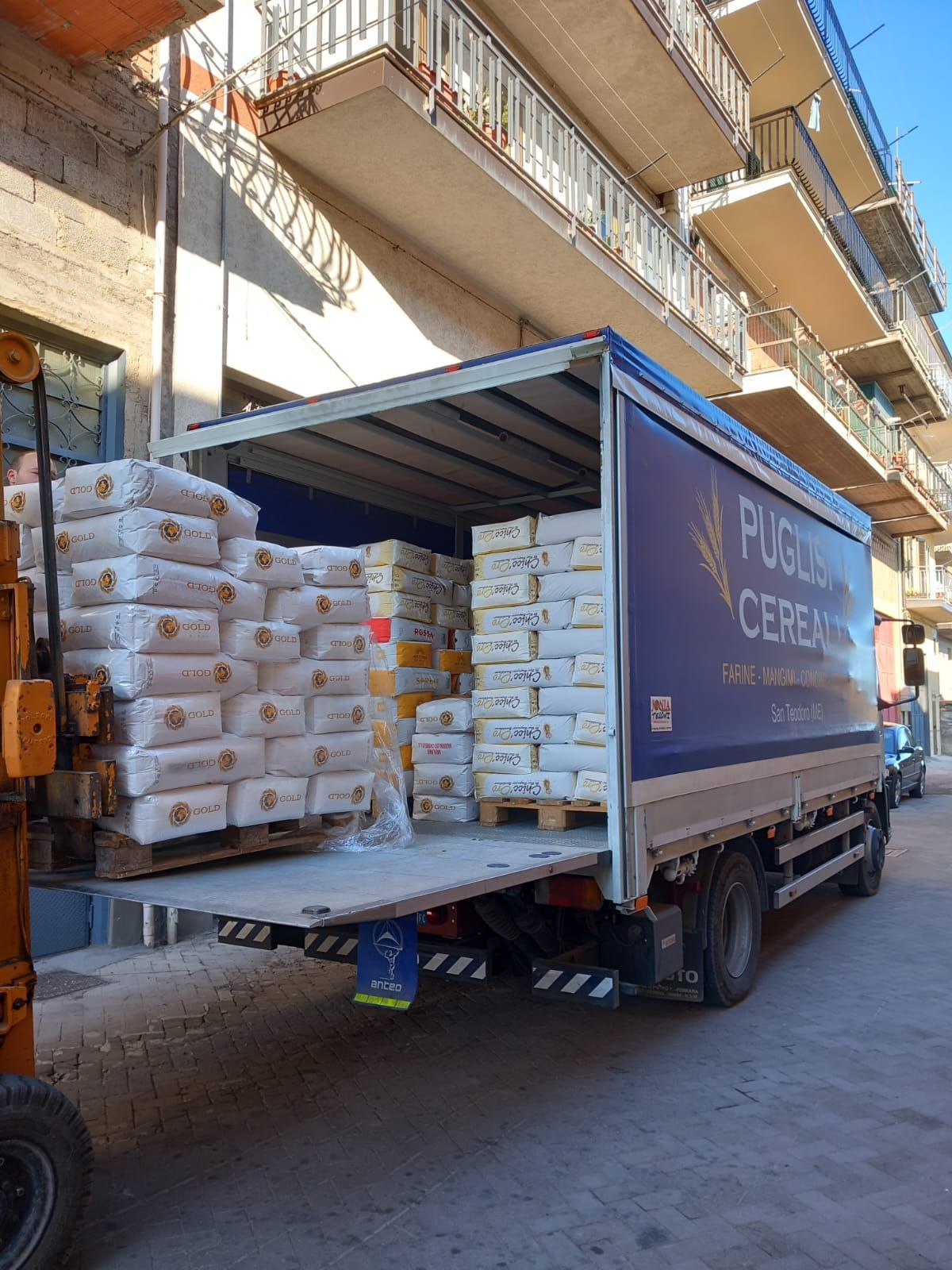 Un camion per le consegne parcheggiato in una strada cittadina, con il cassone pieno di sacchi bianchi di grano impilati su pallet di legno.