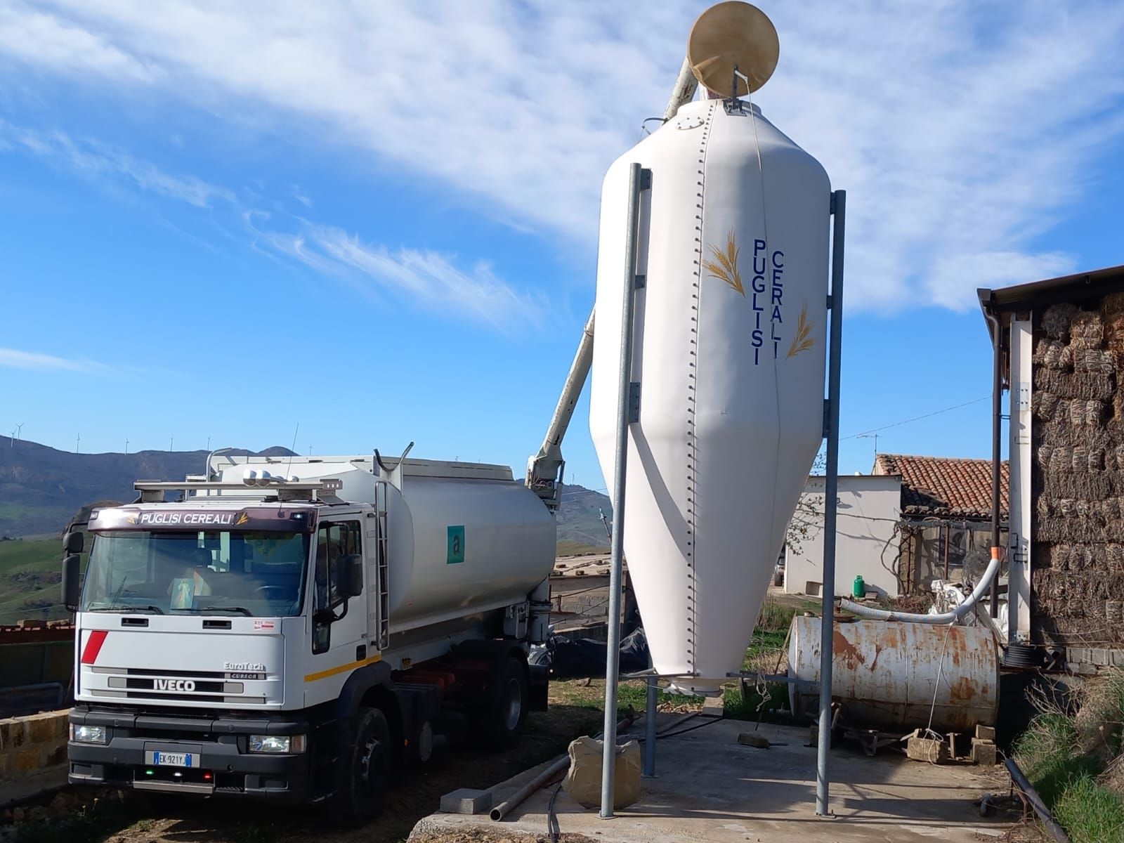 Un'autocisterna bianca utilizza un tubo pneumatico per trasportare il mangime in un grande silo bianco sopraelevato in una fattoria.