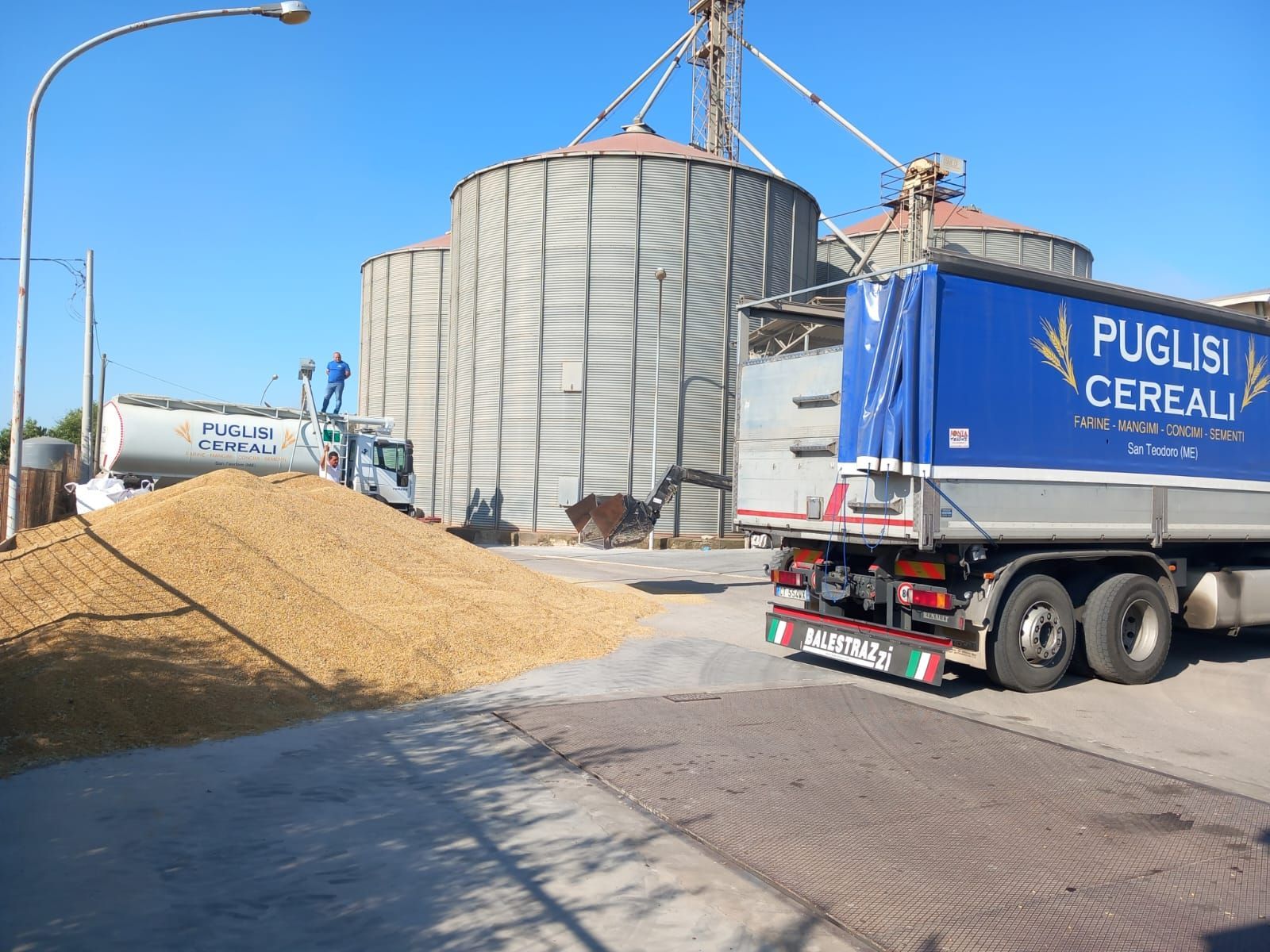 In una giornata di sole, un grande cumulo di grano si trova vicino a un camion e ai silos della Puglisi Cereali presso un impianto di carico.