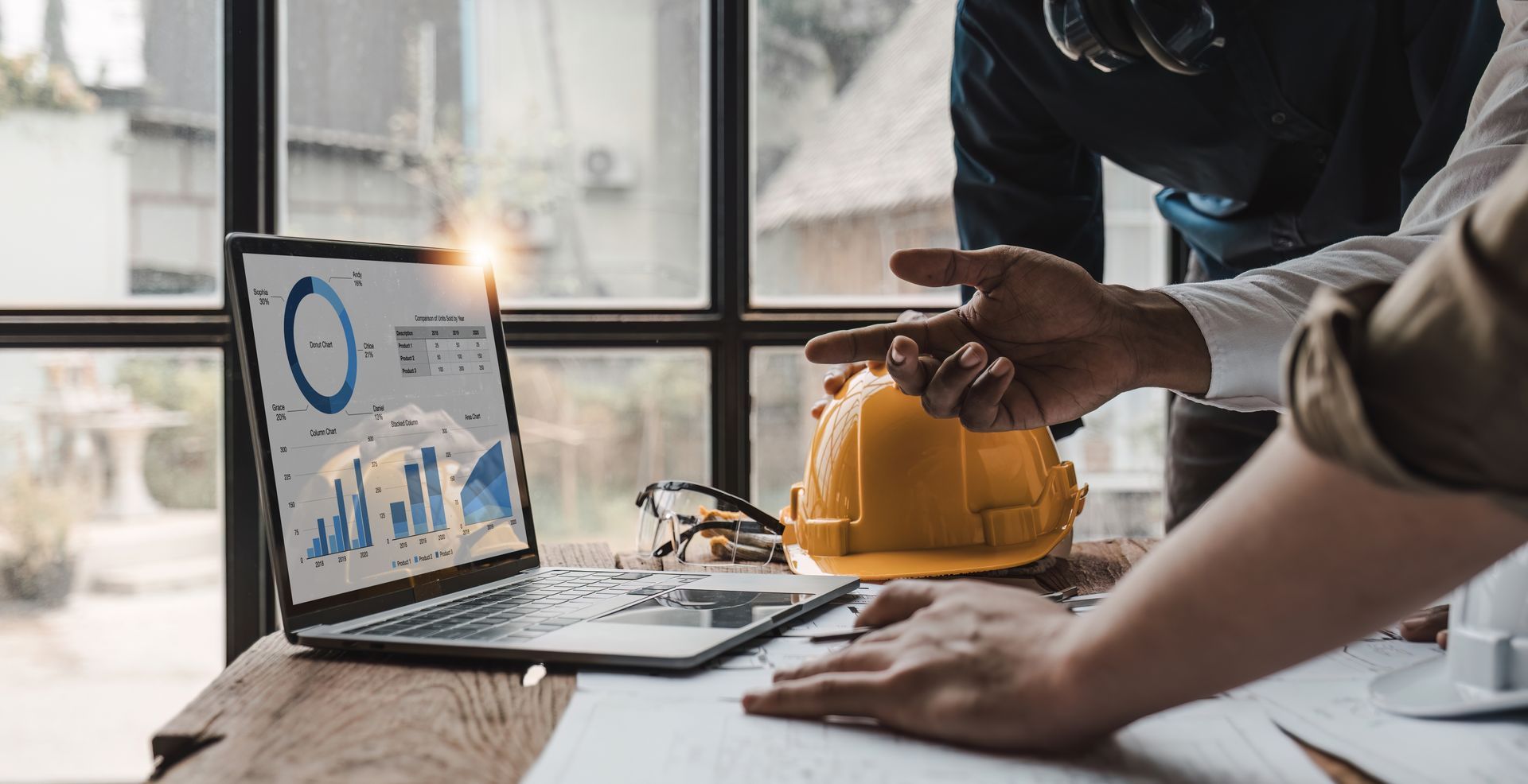 Two people review data on a laptop screen and blueprints at a wooden table next to a yellow hard hat.