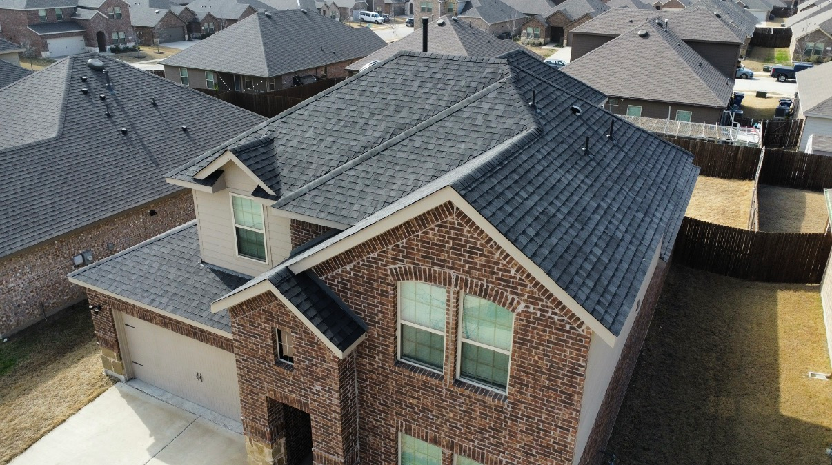 An aerial view of a two-story brick suburban house with a dark shingled roof, surrounded by similar residential homes.