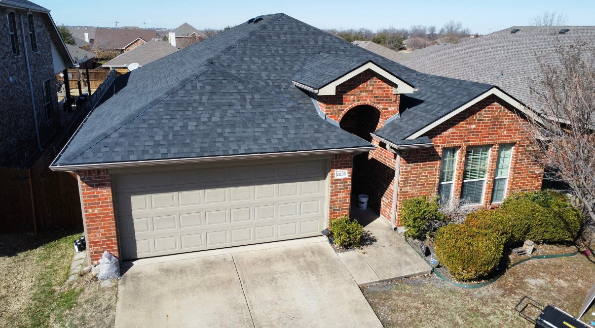 A brick house with a dark shingled roof, a tan garage door, and a small front garden, viewed from above.