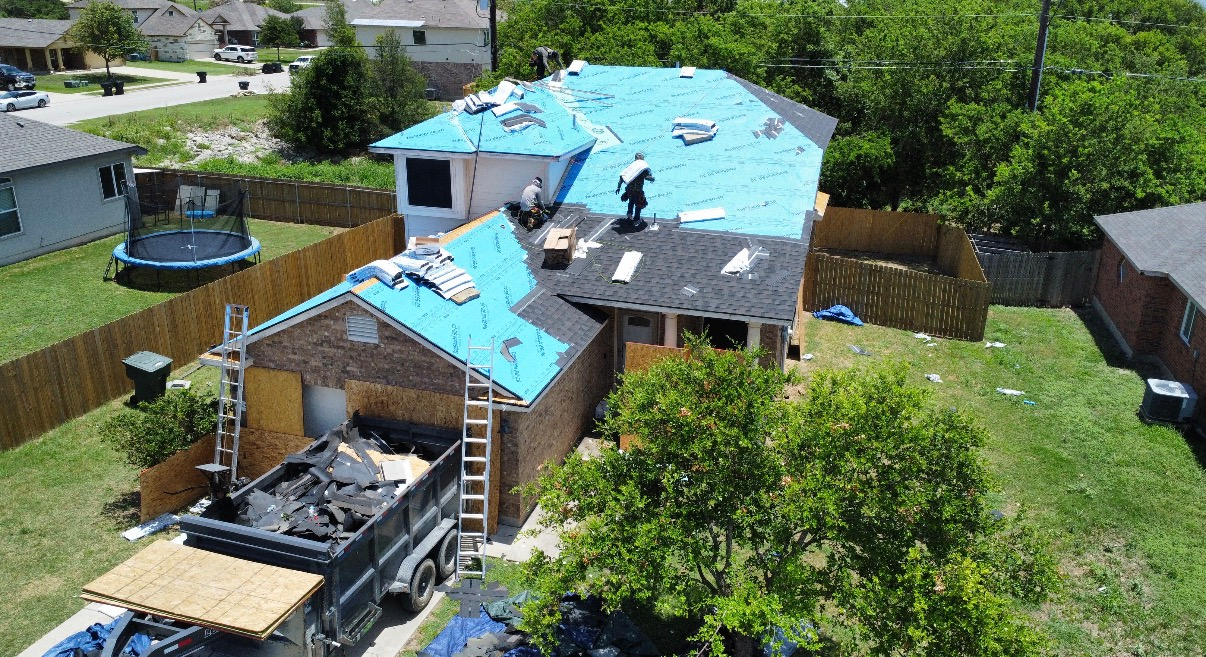 Aerial view of a residential roof under construction with blue synthetic underlayment, black felt, and a dumpster below.