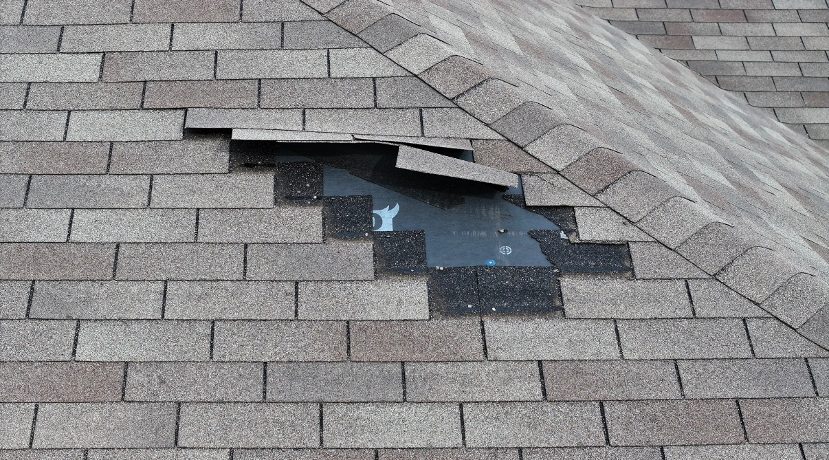 A close-up of a damaged roof with missing gray shingles, revealing the dark underlayment of the roof deck.