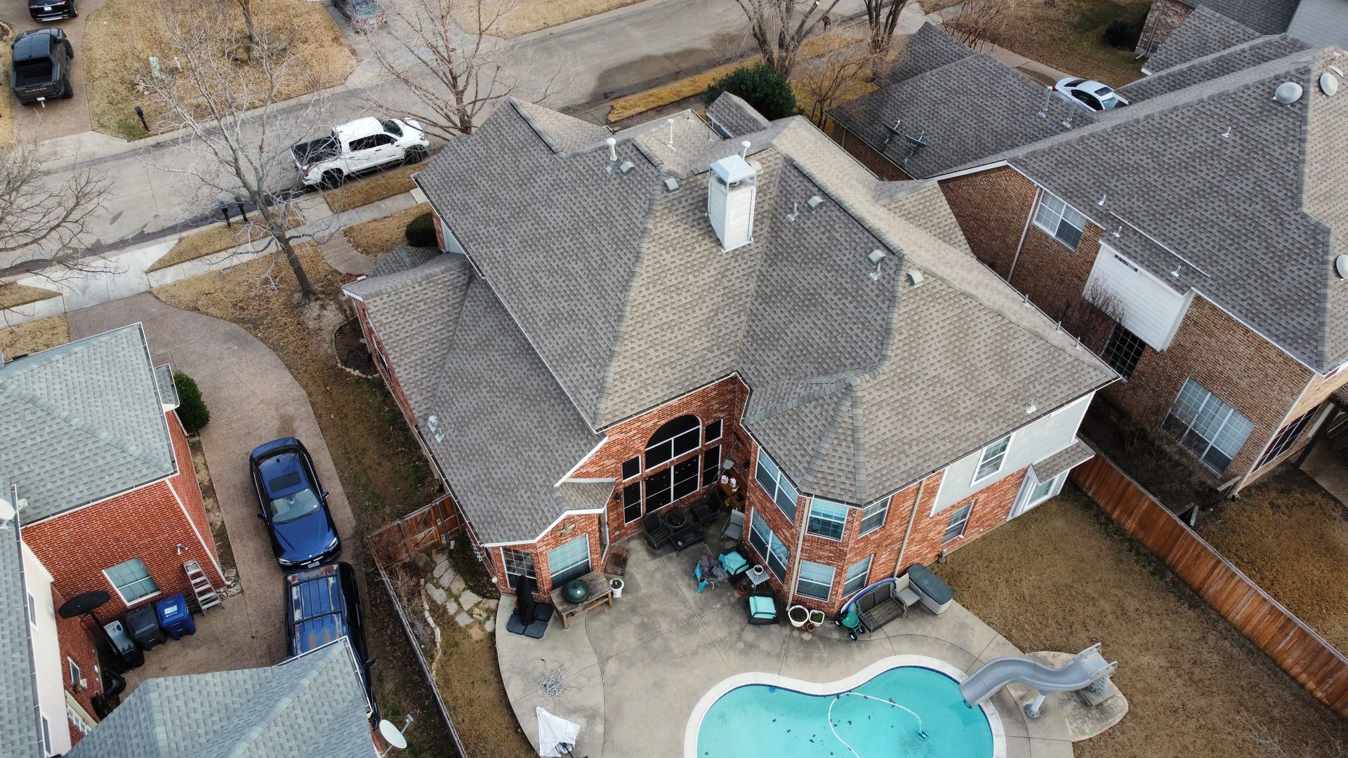 An aerial view of a two-story brick suburban house with a gray roof, a swimming pool, and a driveway with parked cars.
