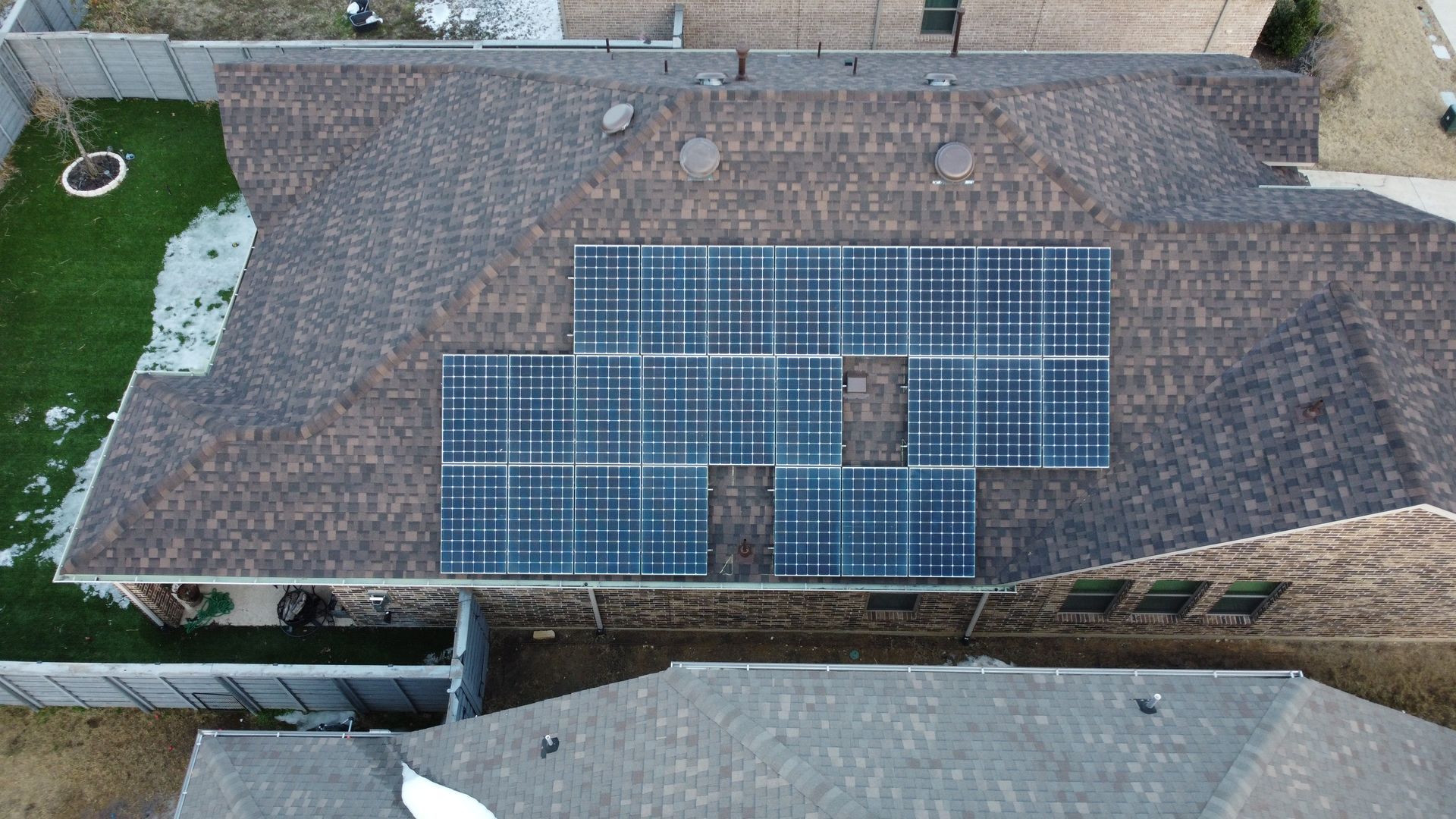 Overhead view of a house with solar panels on a brown shingle roof; the house is surrounded by green grass and other homes.