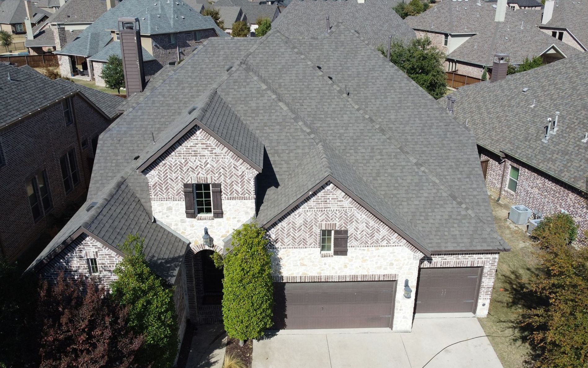 An aerial view of a two-story suburban house with a grey shingled roof, light stone facade, and a three-car garage.