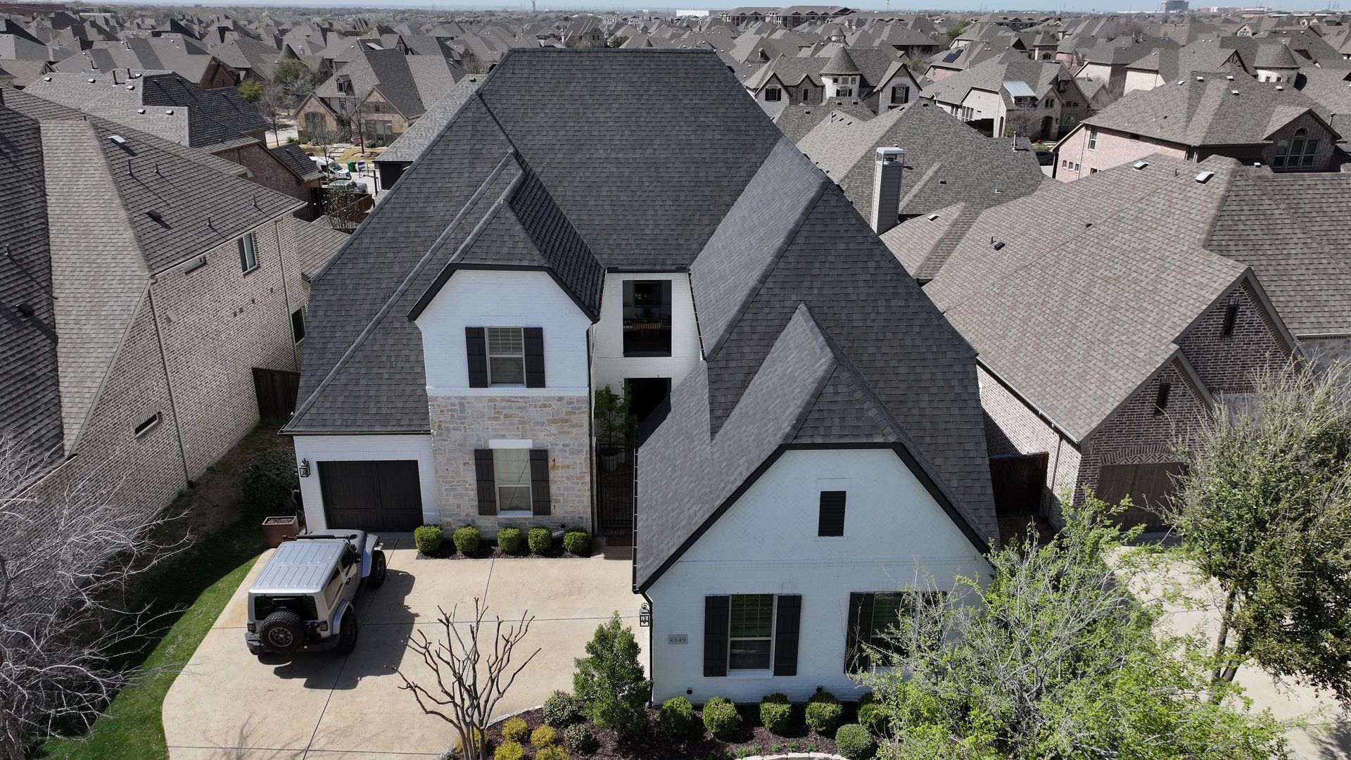 Aerial view of a white and gray house with black roof, parked car, and surrounding suburban homes.
