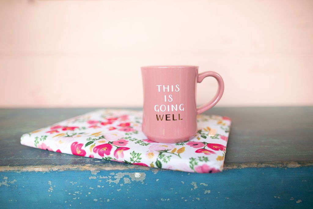 A pink mug and a floral portfolio on a blue table. Mug inscription is humorous This Is Going Well.