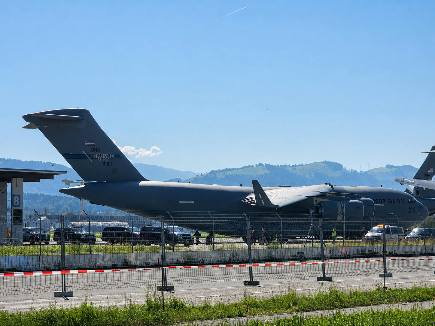 Avion cargo militaire stationné sur une piste derrière une clôture sous un ciel bleu dégagé