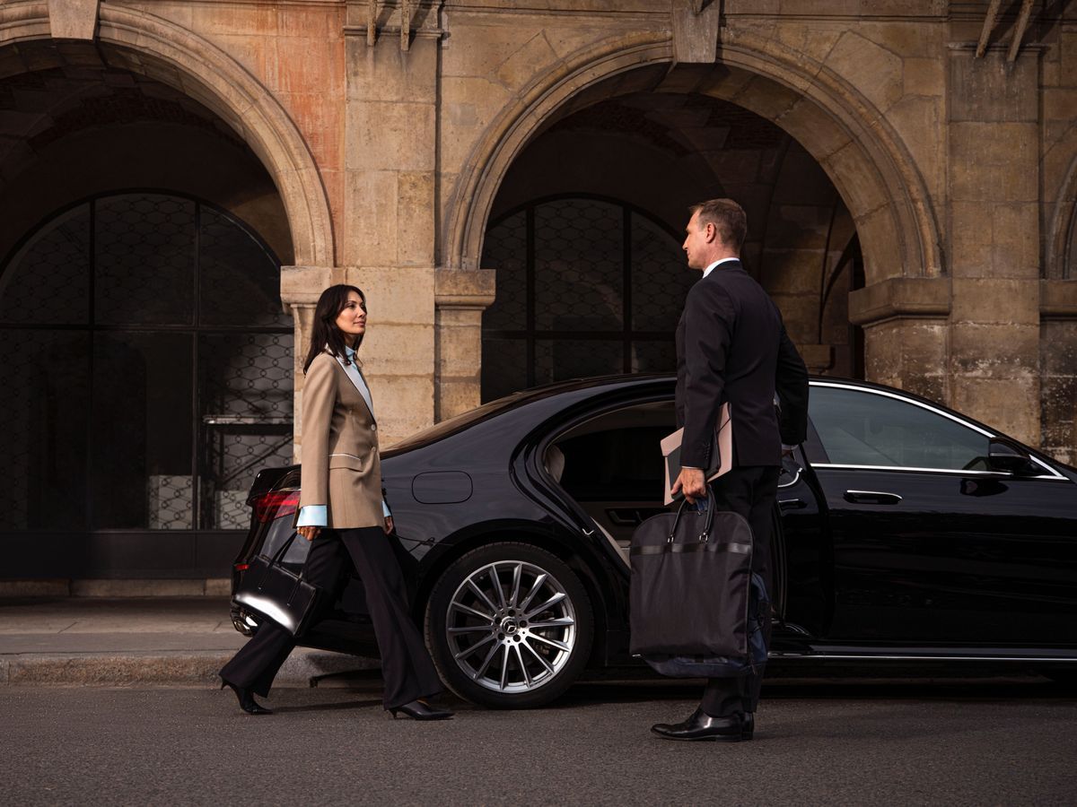 Une femme avec des bagages s'approche d'une voiture noire tandis qu'un chauffeur se tient à proximité, sous des arches de pierre.