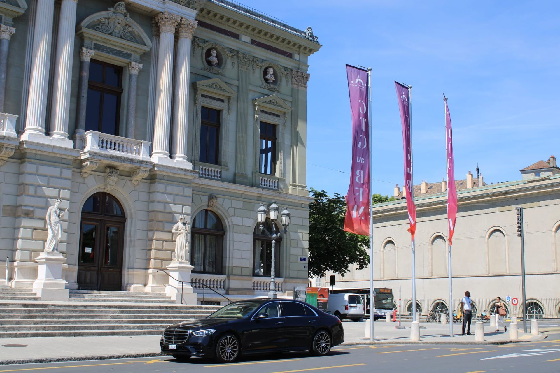 Bâtiment en pierre orné de colonnes et de bannières roses, situé dans une rue ensoleillée, une voiture noire passe à proximité.