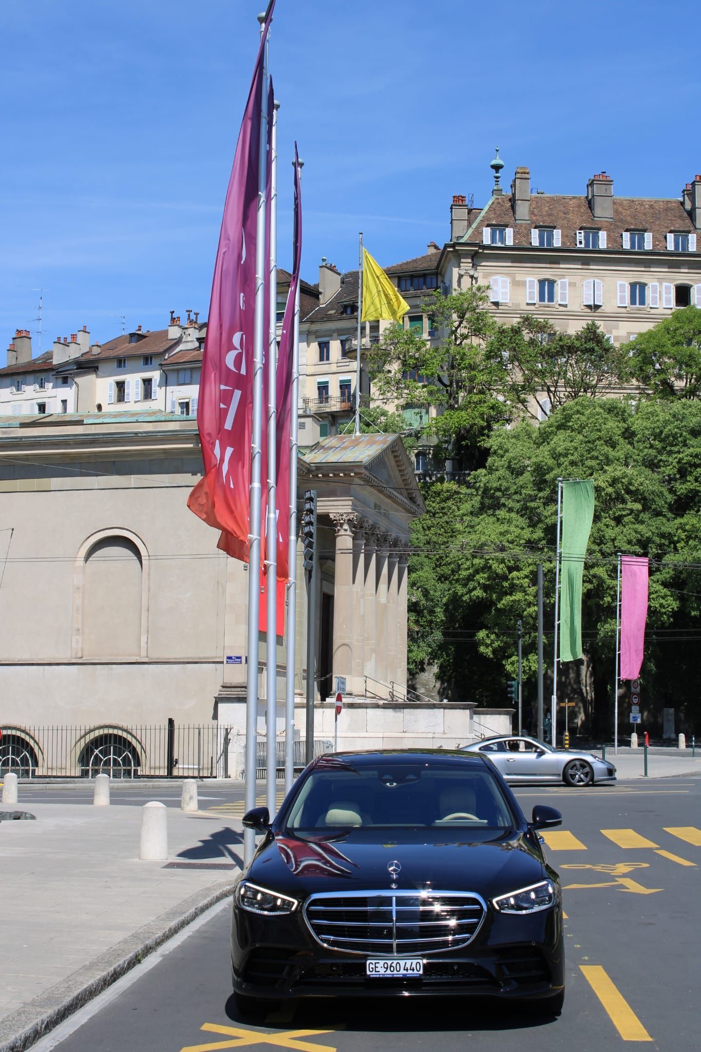 Voiture de luxe noire dans une rue bordée de drapeaux colorés, avec des bâtiments historiques sous un ciel bleu.
