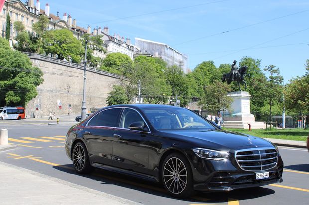 Une berline de luxe noire circule dans une rue ensoleillée, près de murs de pierre et d'arbres, en milieu urbain.